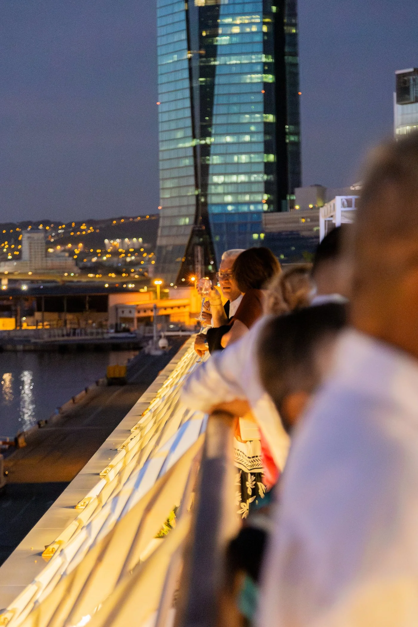 Groupe de personnes assises le long d'une rambarde au bord d'un quai ou d'une promenade, avec un bâtiment moderne en verre au coucher du soleil ou en soirée, évoquant une ambiance urbaine et détendue.