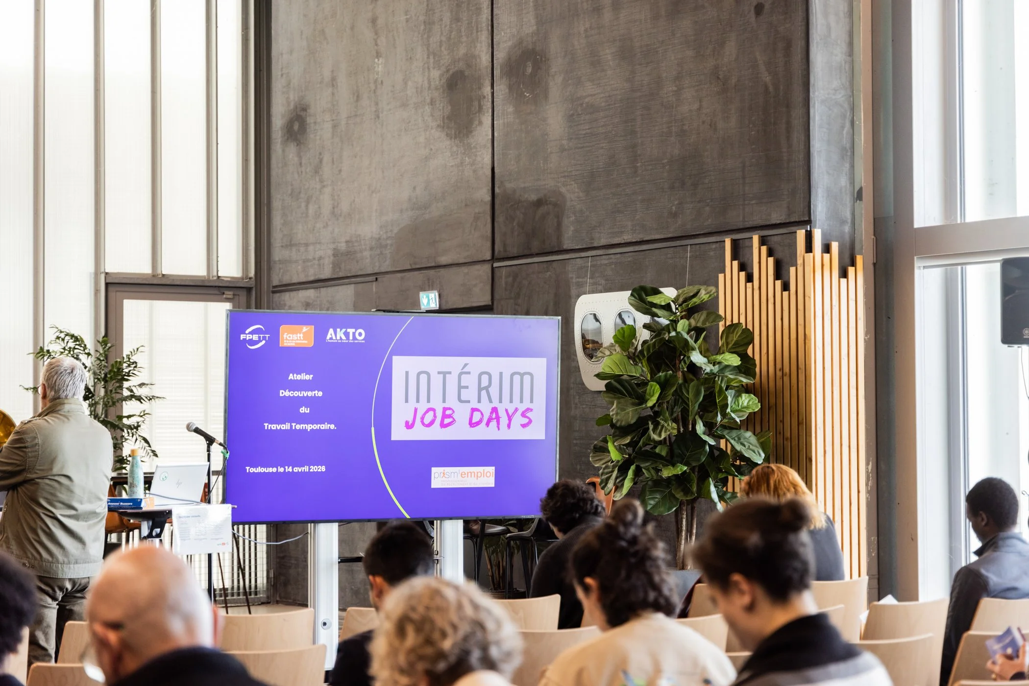 Une salle de conférence avec un grand écran violet affichant un événement intitulé 'INTÉRIM JOB DAYS' organisé par Prisme Emploi le 14 avril 2026 à Toulouse, avec des personnes assises en train d'assister à la présentation.