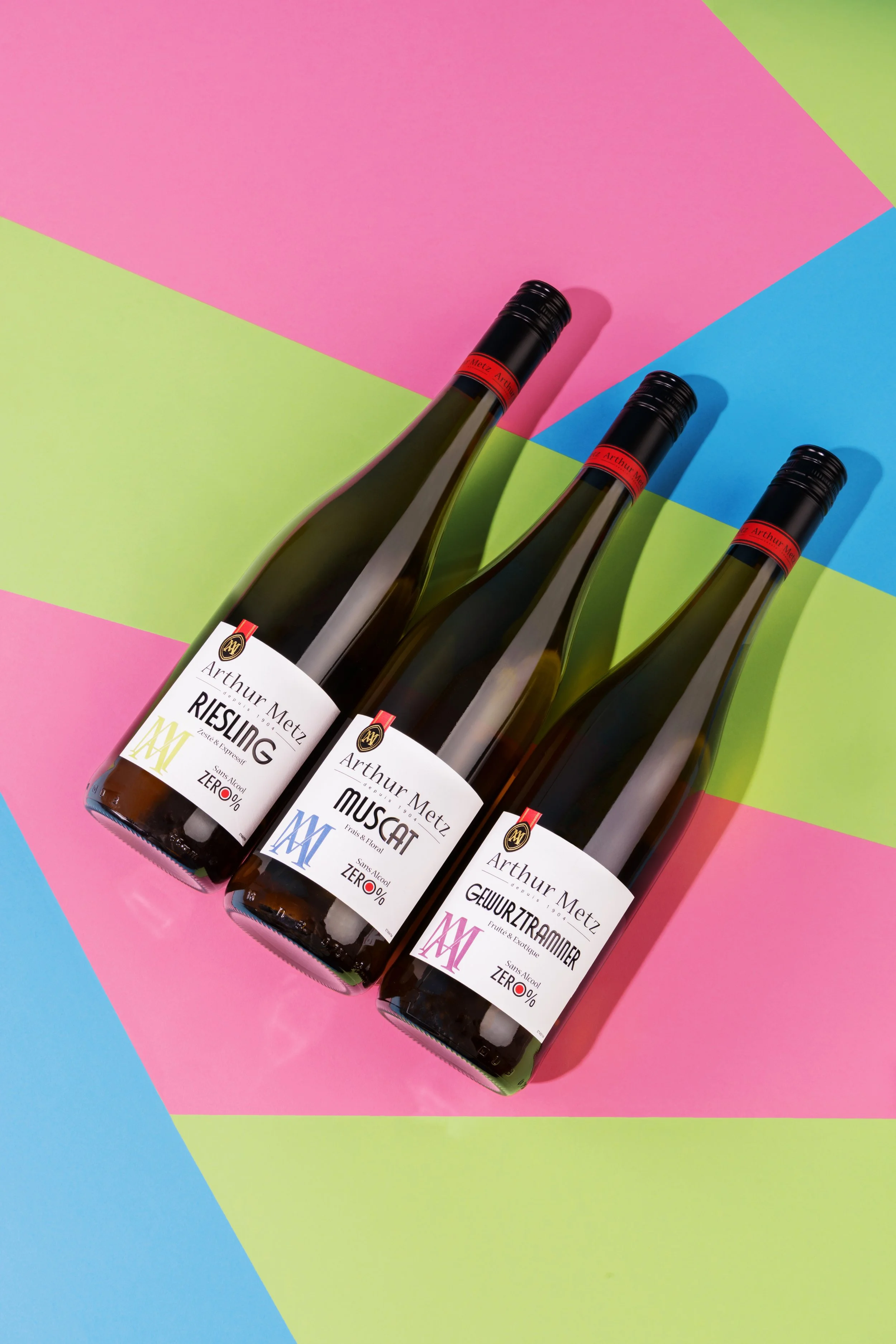Trois bouteilles de vin blanc de marque Arthur Metz posées sur un fond coloré en blocs de rose, vert, bleu et jaune.