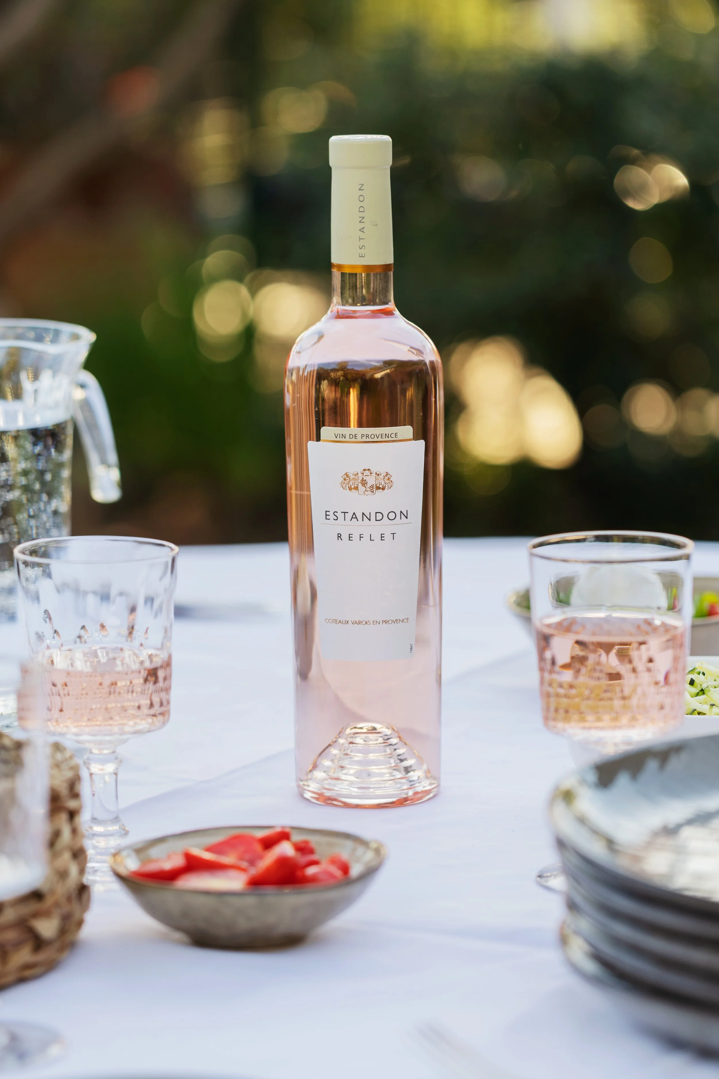 Une bouteille de vin rosé Estandon Reflex au centre d'une table, entourée de verres à vin remplis de rosé, avec des plats de nourriture et un fond flou de nature en plein air.