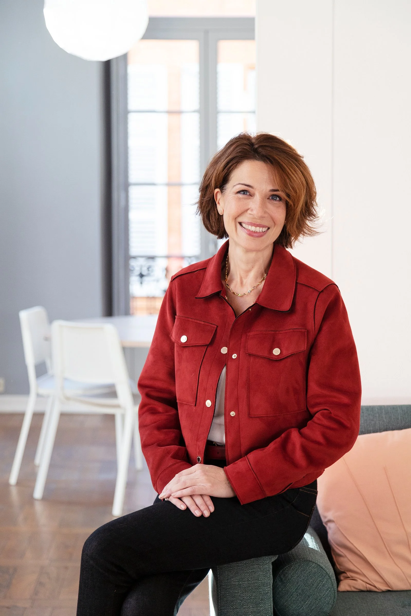 Une femme souriante assise dans un intérieur lumineux avec des fenêtres derrière elle, portant une veste rouge et un chemisier blanc, avec d'autres meubles et chaises dans la pièce.