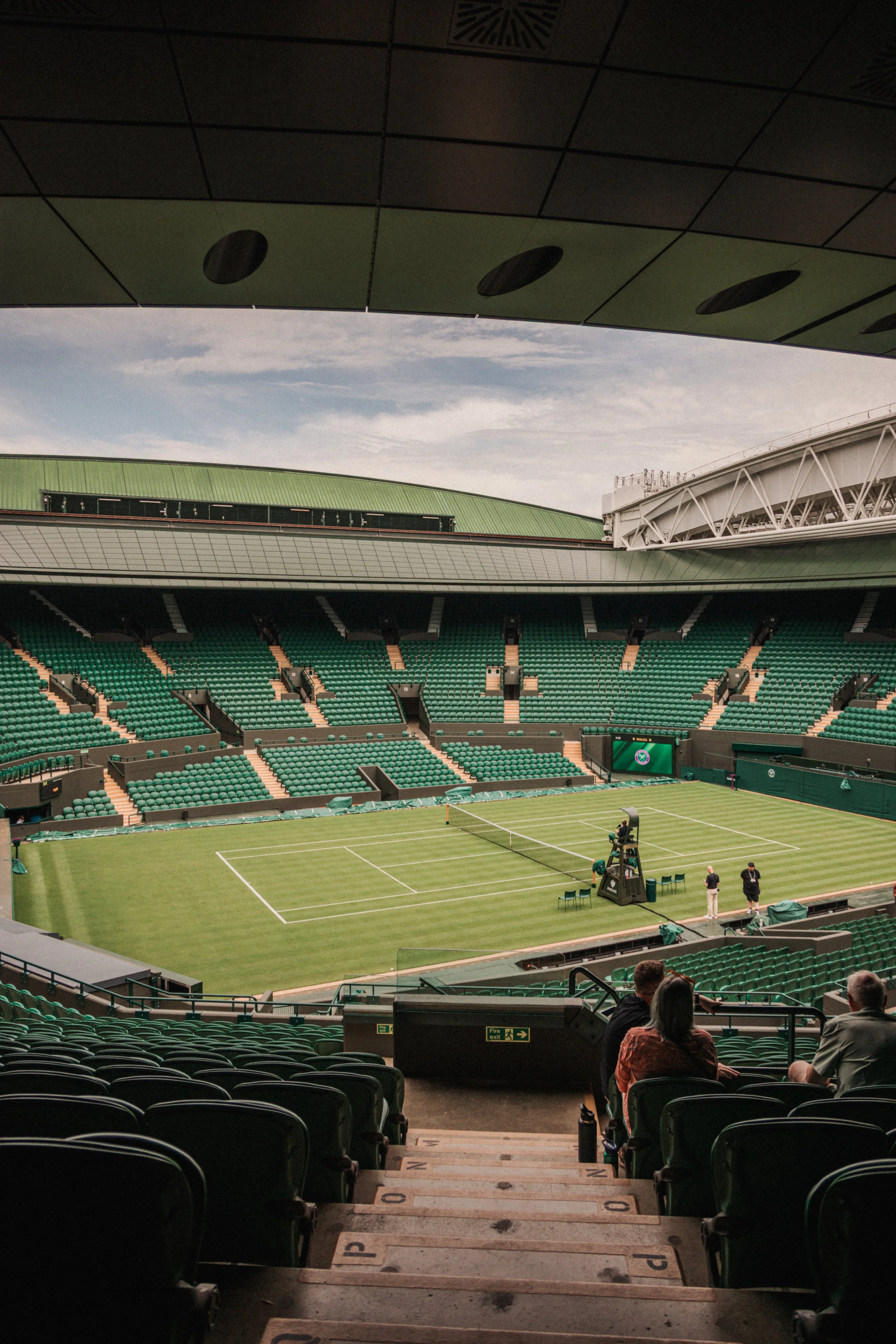 Vue intérieure d'un stade de tennis avec un court en gazon vert, des sièges verts et quelques personnes en préparation pour un match.