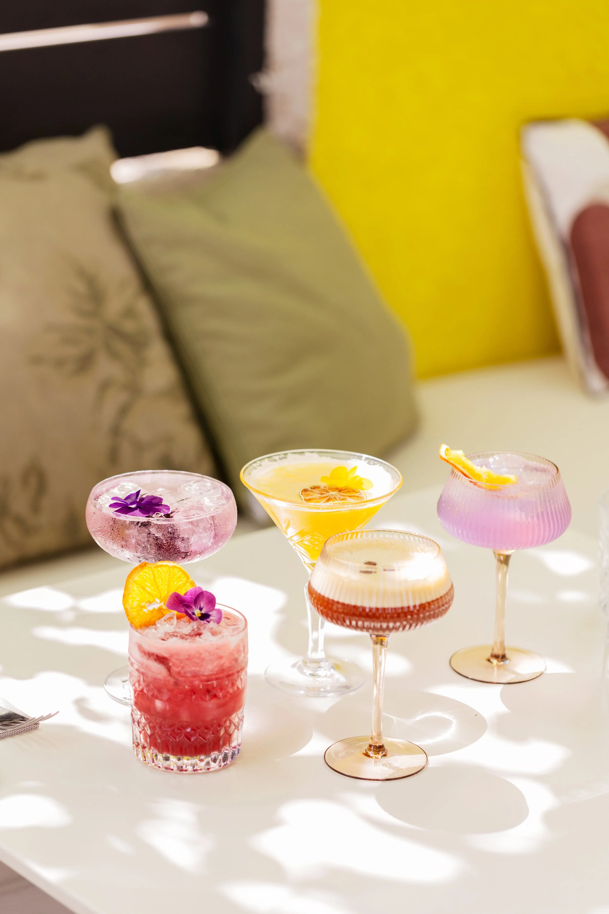 Une table avec plusieurs cocktails colorés décorés de fleurs, de tranches d'agrumes et de garnitures variées, sous un éclairage doux avec un fond de coussins et d'un mur jaune.