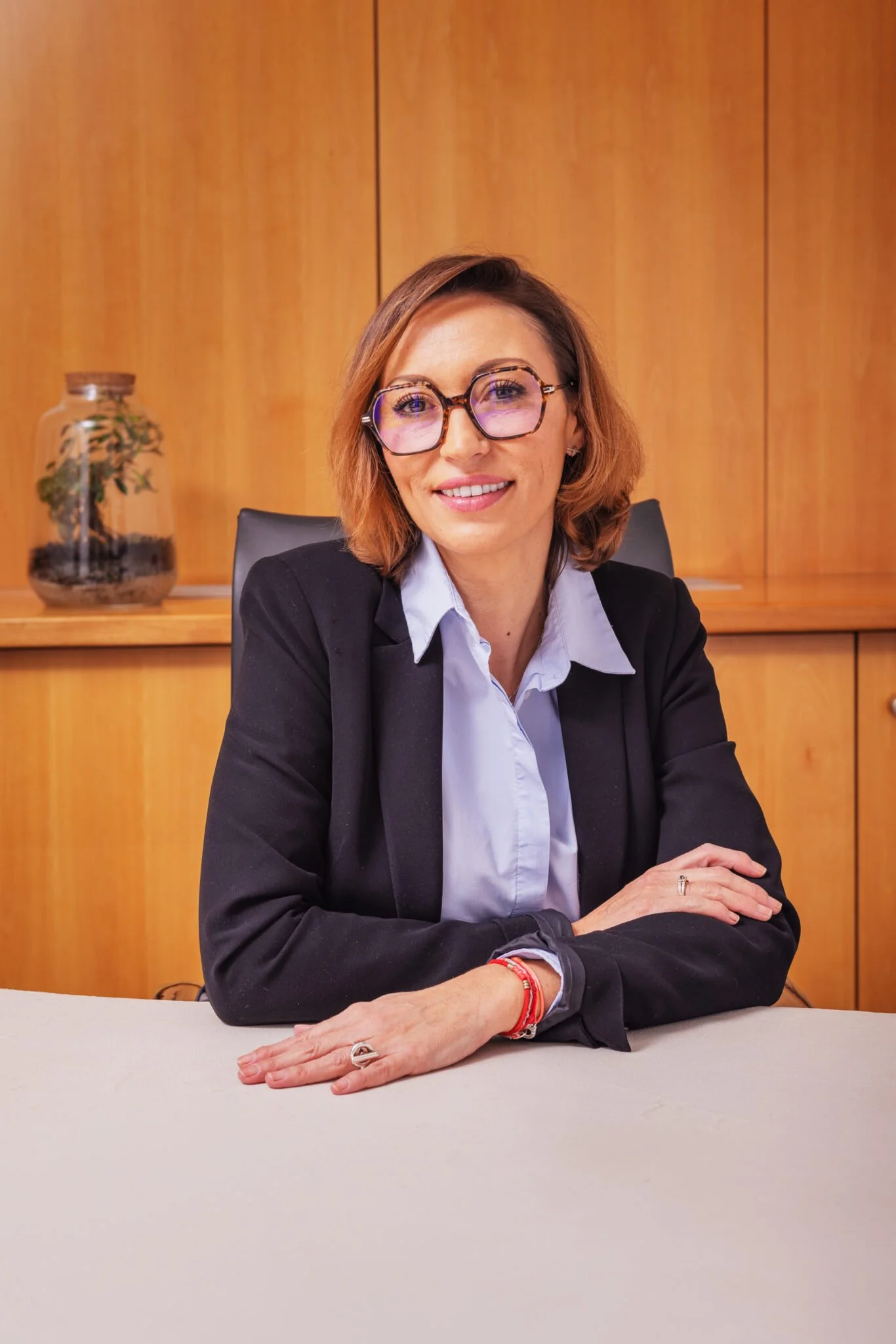 Une femme assise à une table, souriant, portant des lunettes, un blazer noir et une chemise blanche, avec un fond en bois.