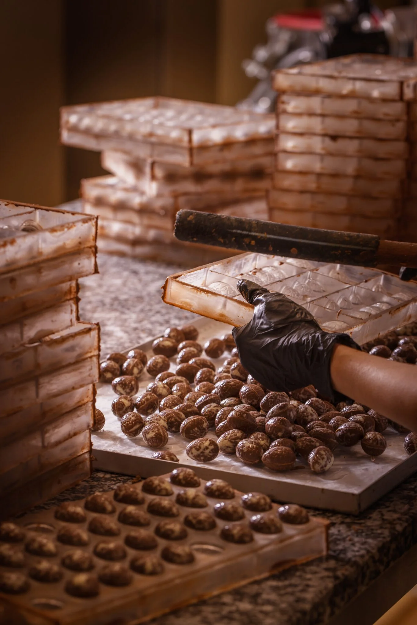 Une personne portant un gant noir manipule des œufs de chocolat dans une boîte en plastique. Plusieurs boîtes en plastique remplies d'œufs de chocolat colorés sont empilées en arrière-plan.