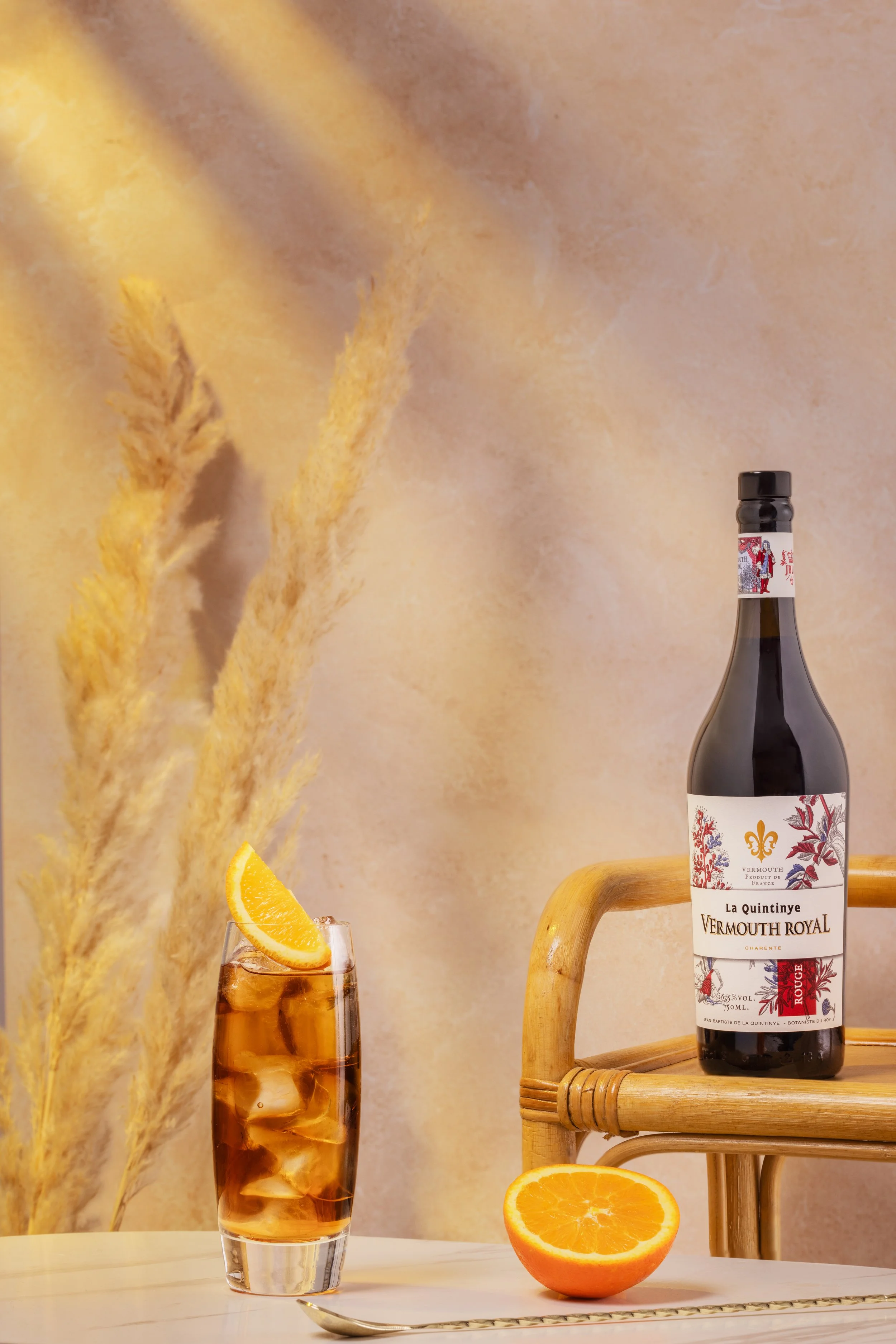 Une bouteille de vermouth Royal La Quintinye avec un verre rempli de boisson glacée garnie d'une tranche d'orange, sur une table blanche avec des tranches d'orange et une cuillère, devant un arrière-plan de décoration en beige et des plantes séchées