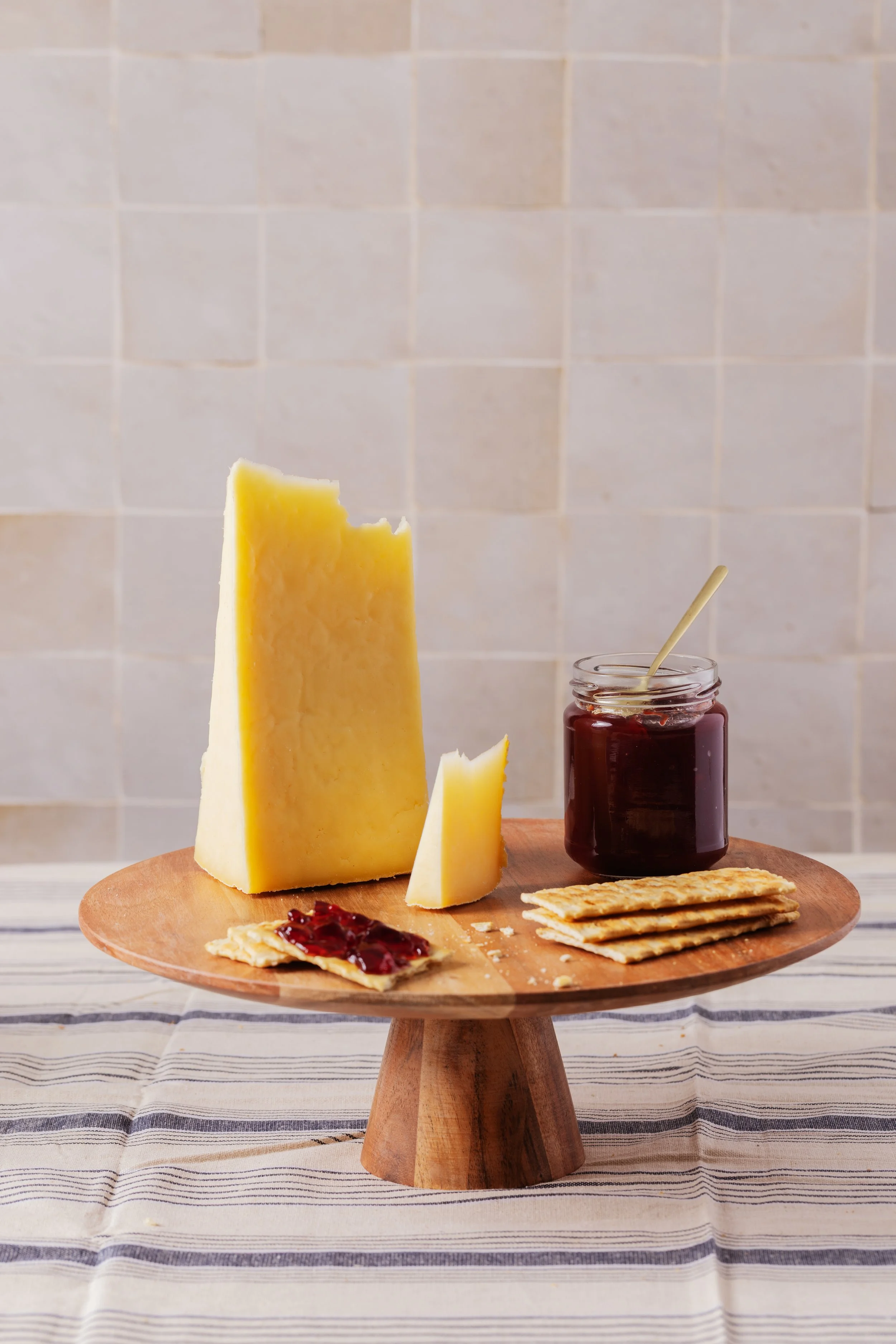 Fromage jaune à pâte dure placé sur un plateau en bois, avec des crackers, confiture de fruits rouges dans un petit bol, et une jarre de confiture de fruits rouges avec une cuillère en bois, sur une table recouverte d'une nappe rayée.
