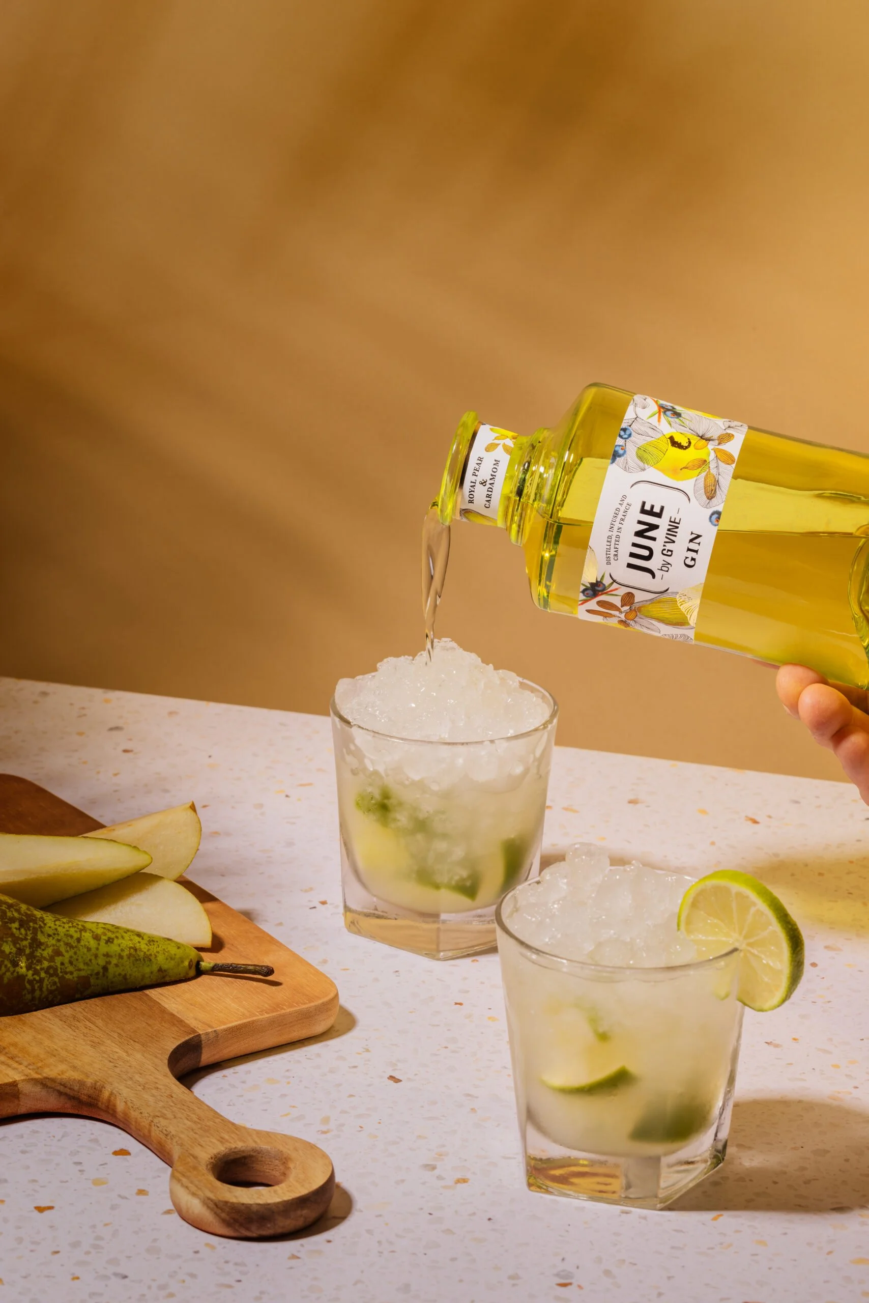 Groupe de trois verres avec cocktail au citron vert, gorgé de glace pilée et de rondelles de citron vert, avec une bouteille de gin aromatisé au poire et cardamome en train d'être versée.