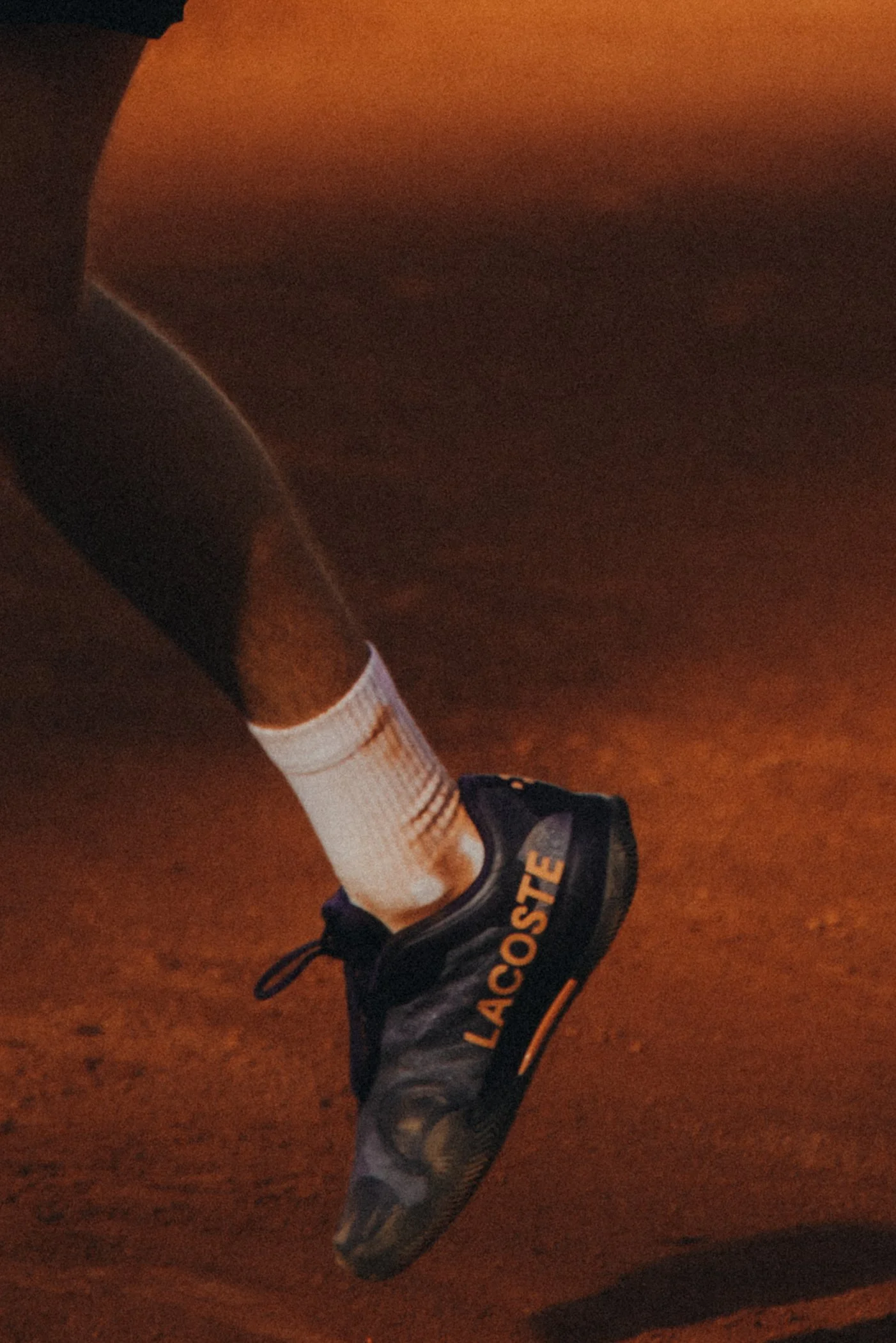 Une chaussure de course noire avec le mot "LACOSTE" en orange, une chaussette blanche avec des rayures orange et rouges, et une jambe chaussée d'un pantalon gris camouflage, sur un sol en terre