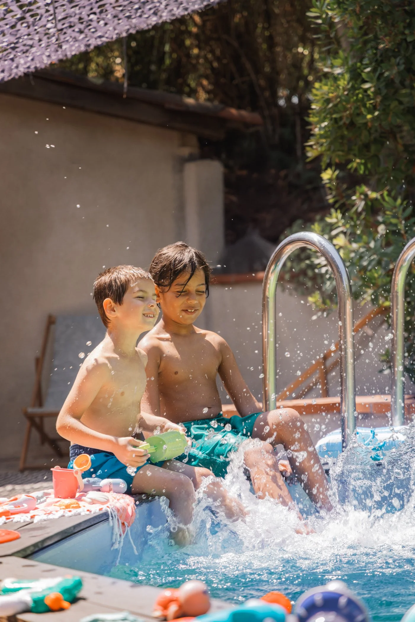 Deux enfants jouent avec de l'eau dans une piscine extérieure, assis sur le rebord, avec des jouets de plage autour d'eux, sous un soleil éclatant, entourés de végétation.