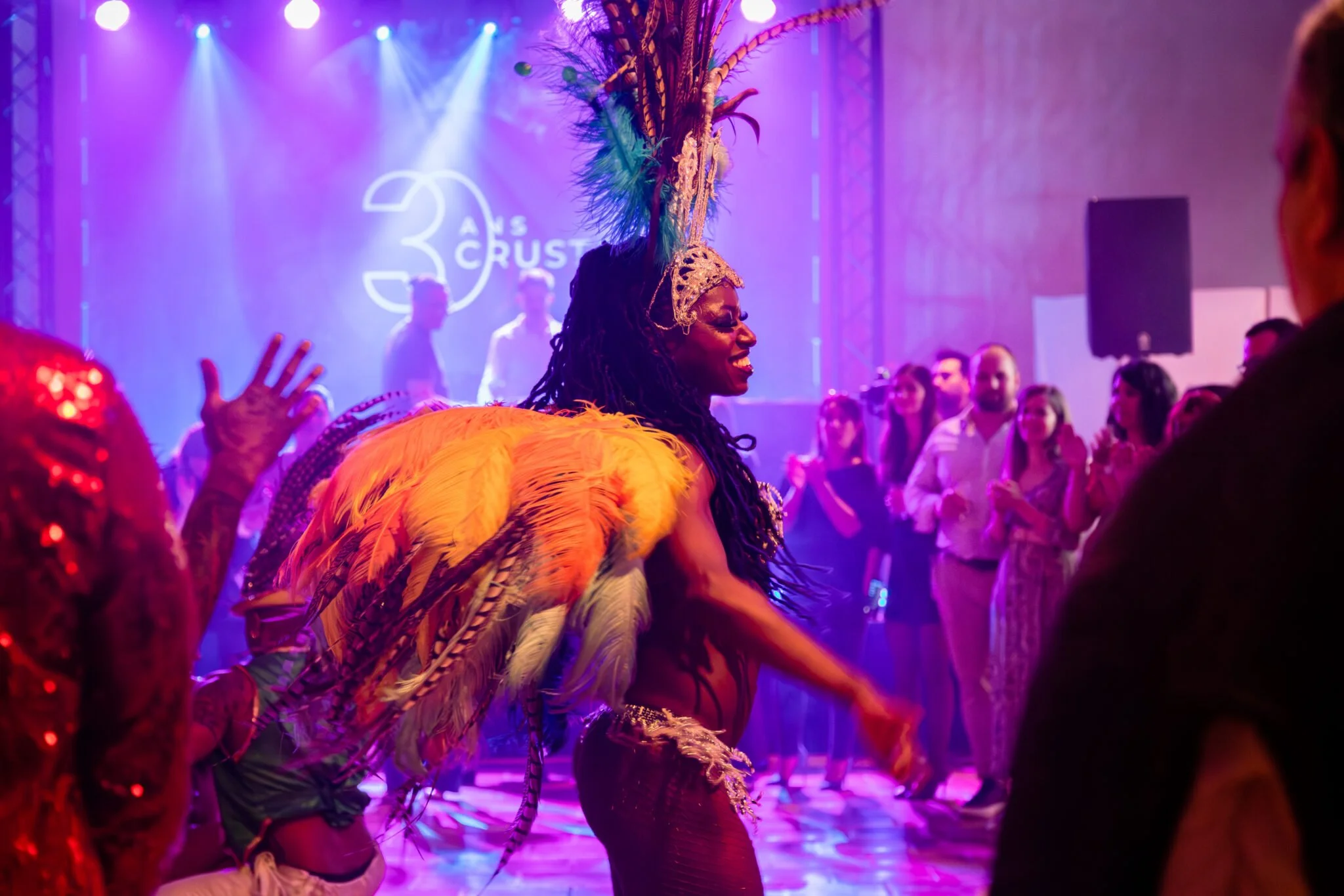 Une femme porte un costume coloré avec des plumes et un grand bonnet à plumes lors d'une performance ou d'une célébration, entourée d'un public dans une ambiance lumineuse violette.