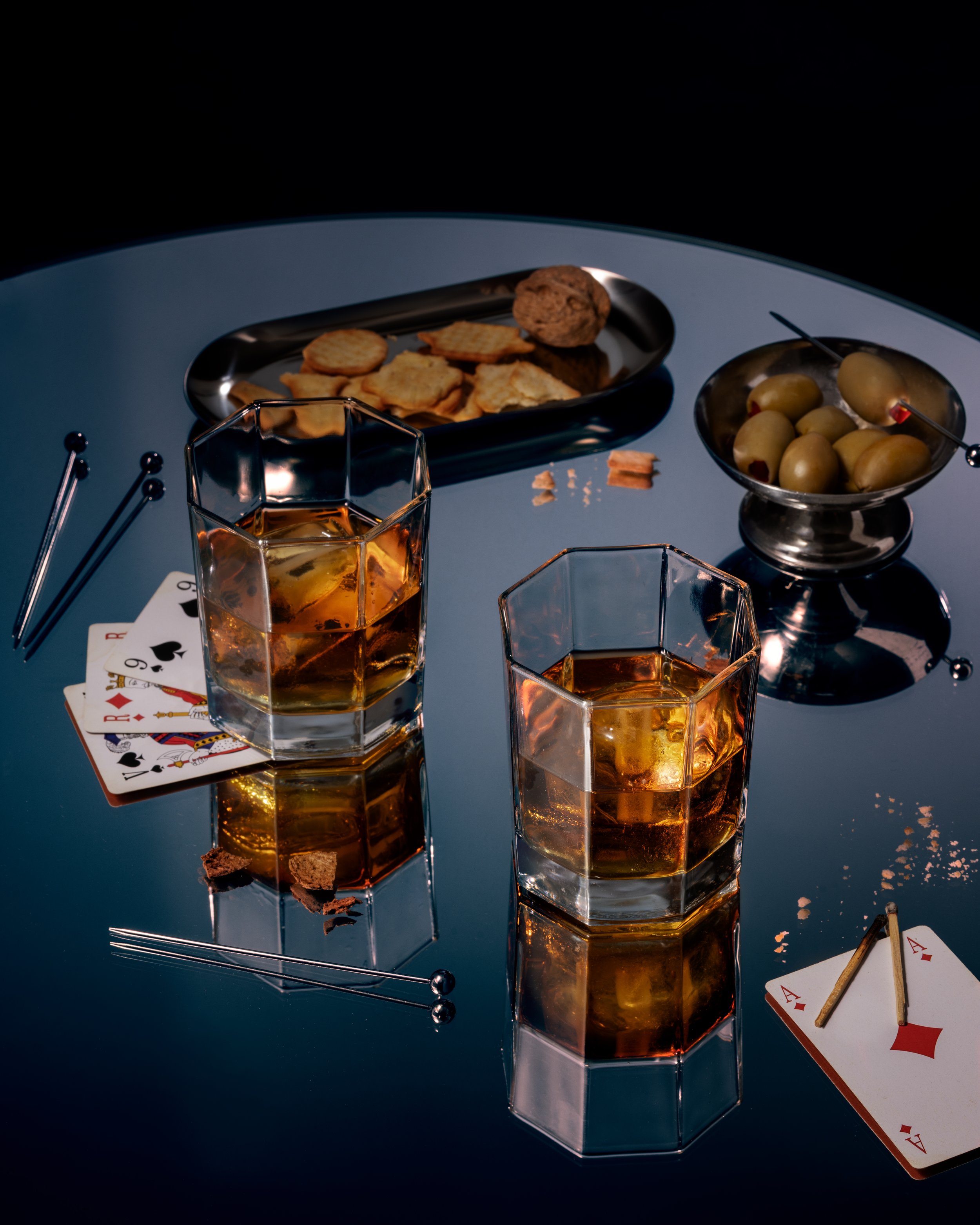 Verre de whisky en verre géométrique sur une table noire, avec des biscuits sur une assiette en métal, une boule de viande ou de pâté, et des chips. Cartes à jouer et bagues à sourcils sur la table.