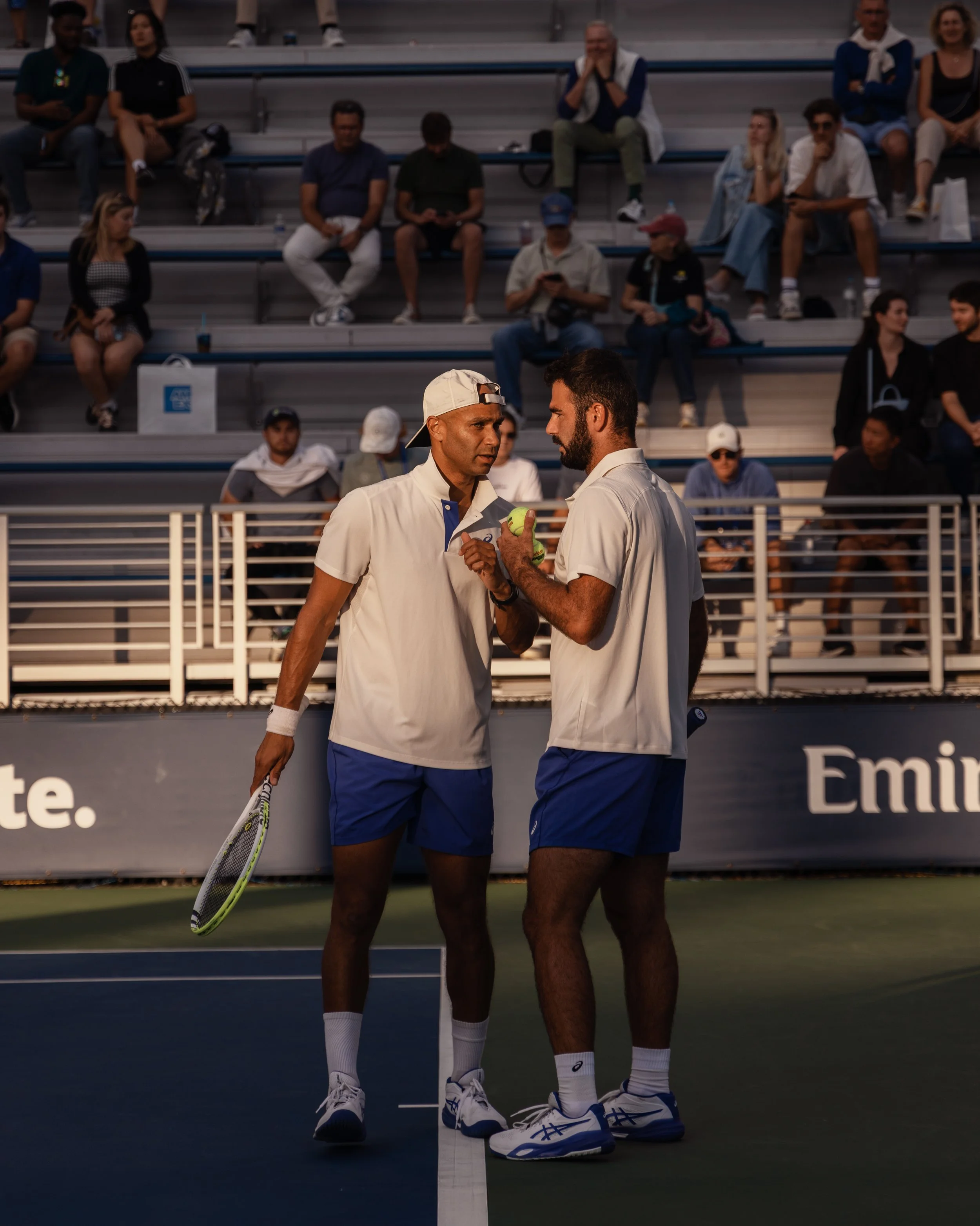 Deux hommes habillés en tenue de tennis, discutant et se serrant la main ou se donnant un accolade sur un court de tennis, avec des spectateurs en arrière-plan.