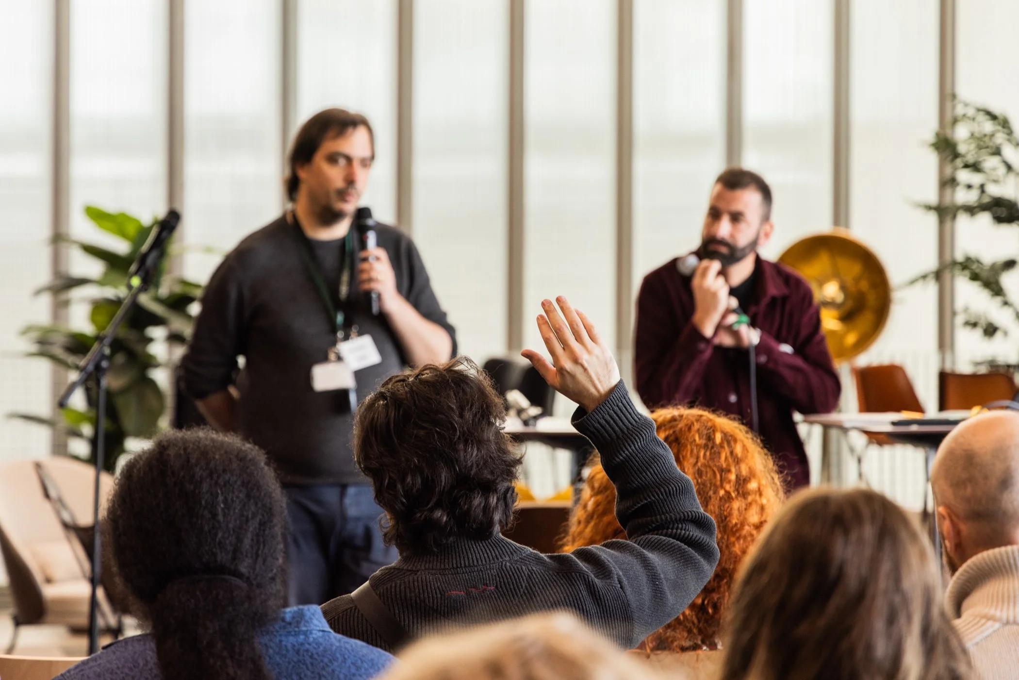 Deux hommes parlant dans un environnement intérieur lors d'une conférence ou d'un atelier, avec un public de personnes qui posent une question, et des plantes en arrière-plan.