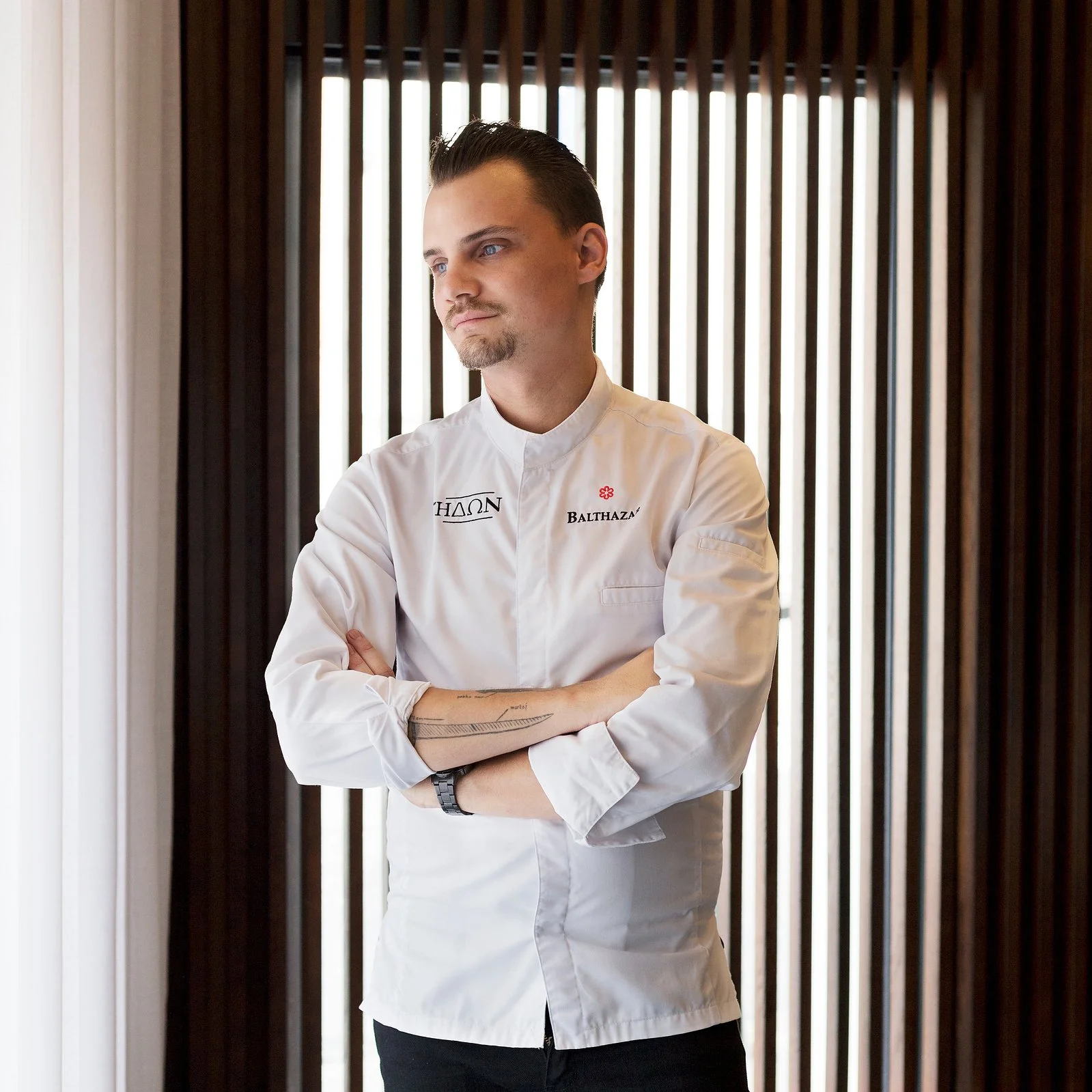 Un homme en uniforme de chef cuisinier, portant une veste blanche avec des inscriptions, croise les bras et regarde pensivement.