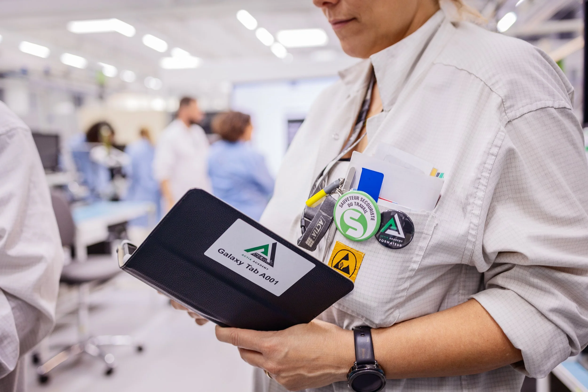 Une femme porte un badge avec le logo de l'académie Galaxy Tab et une mention indiquant le modèle A007. Elle tient une tablette ou un carnet. Elle porte un bracelet noir et une veste blanche. Le fond montre un environnement d'infirmière ou hospitalie