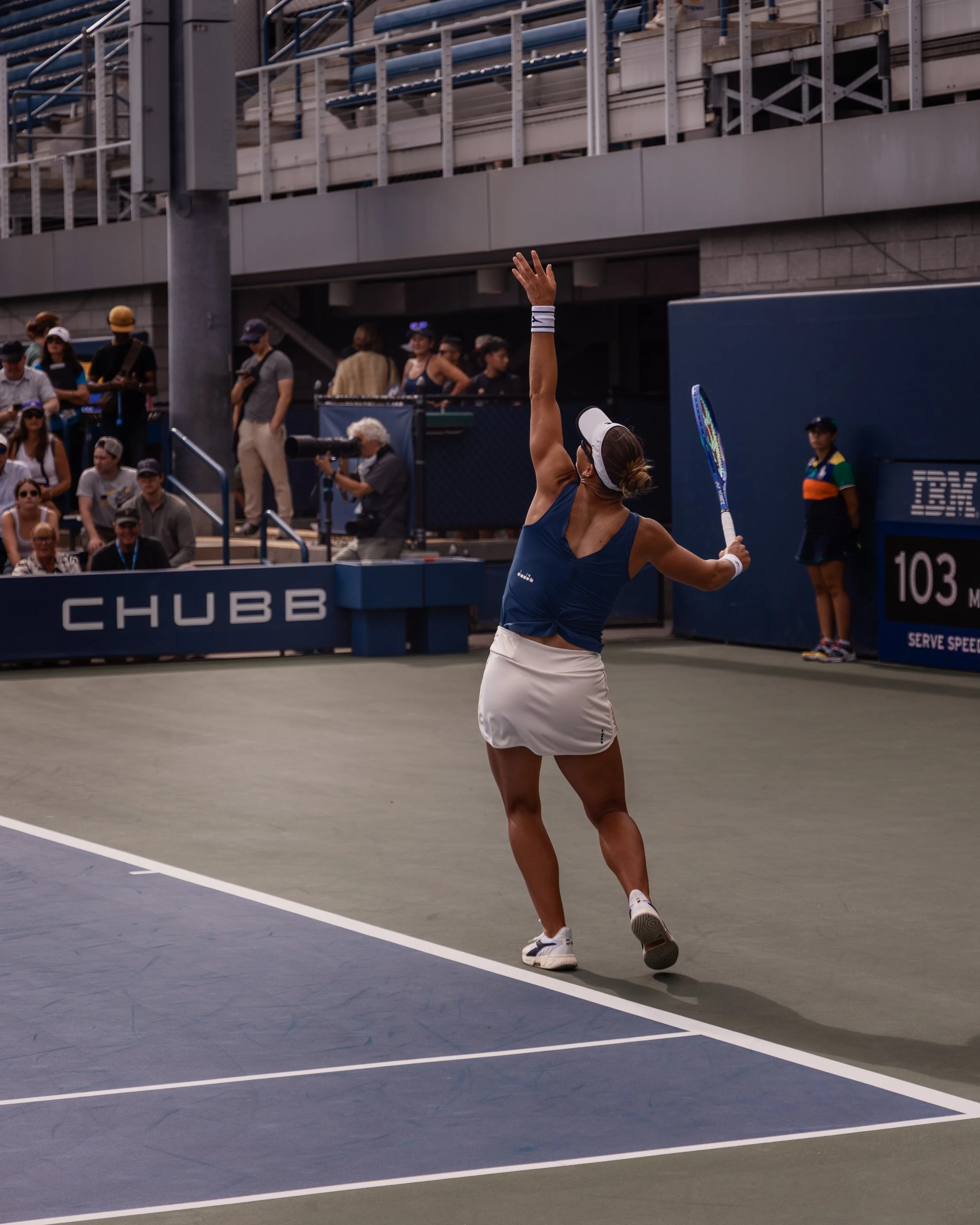 Une femme en tenues de sport blanc et bleu joue au tennis sur un court intérieur, avec une foule de spectateurs en arrière-plan et un homme filmant la scène.
