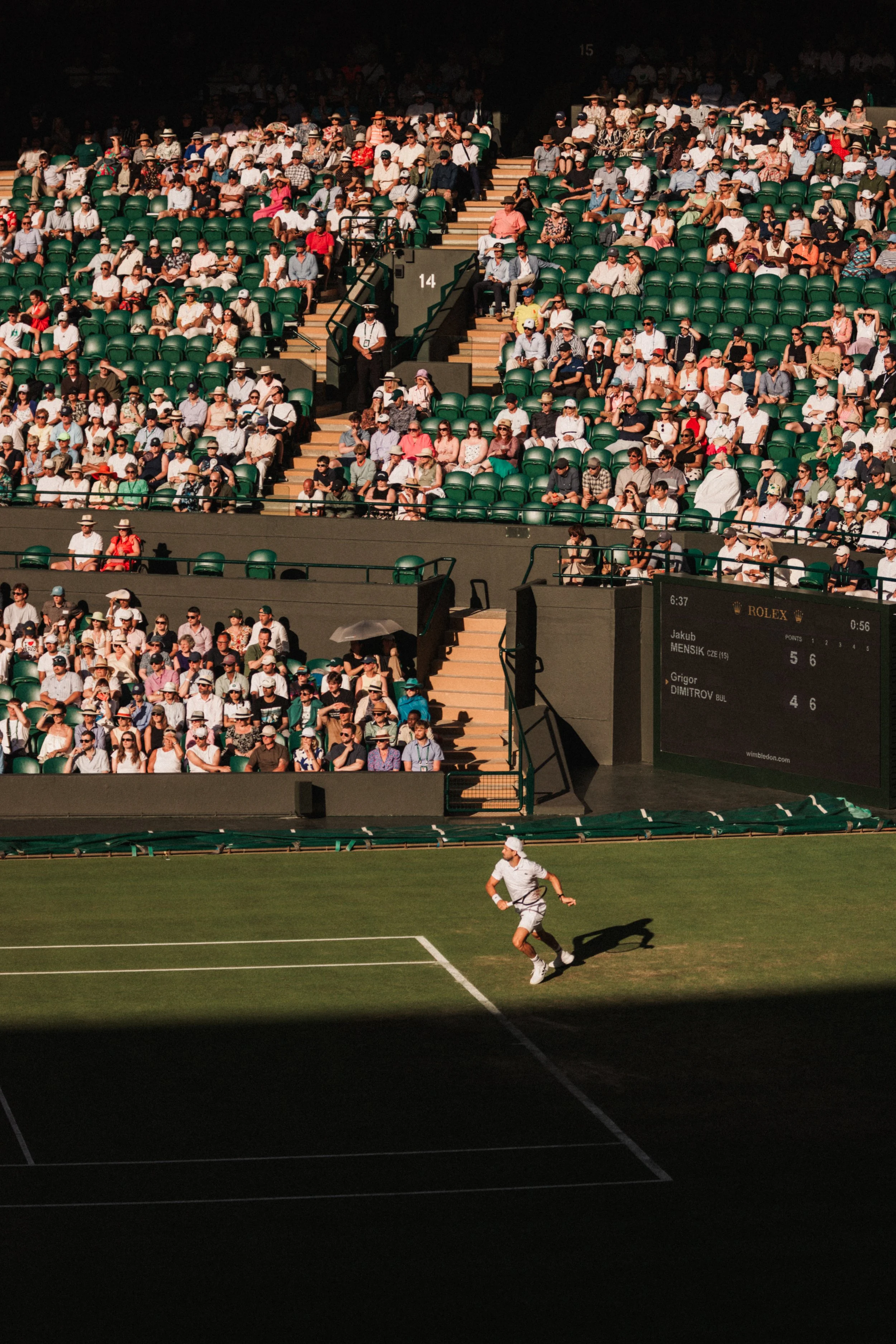 Un joueur de tennis court au sol sur un court en herbe entouré d'un public nombreux assis dans un stade, le public porte des chapeaux et des vêtements légers, le score affiché indique Jakub MENSIK contre Grigor DIMITROV.