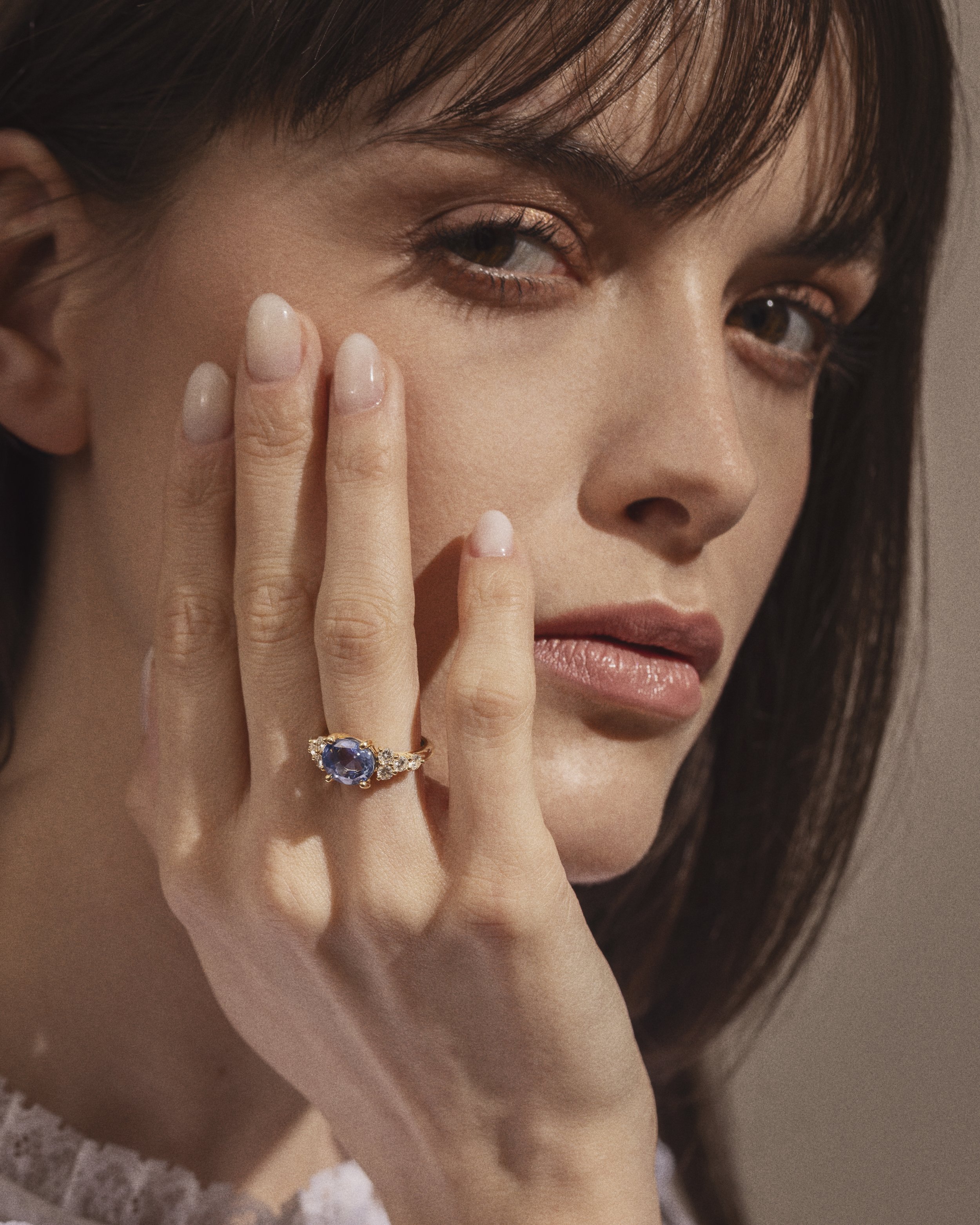 Close-up d'une femme avec des yeux marron, portant un anneau en or avec une pierre bleue sur sa main, avec des ongles couleur nude, près de son visage. Elle a des cheveux bruns et un maquillage naturel.