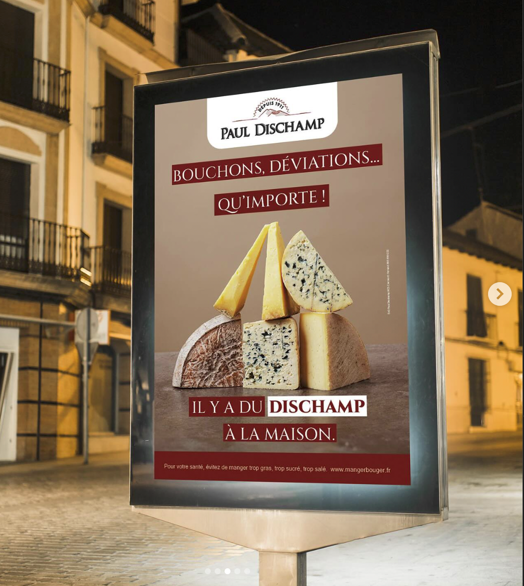 Une affiche publicitaire pour la fromagerie Paul Dischamp, mettant en avant différents types de fromages accompagnée du slogan 'Bouchons, déviations... qu'importe ! Il y a du Dischamp à la maison', avec un fond de bâtiments résidentiels en arrière-plan.