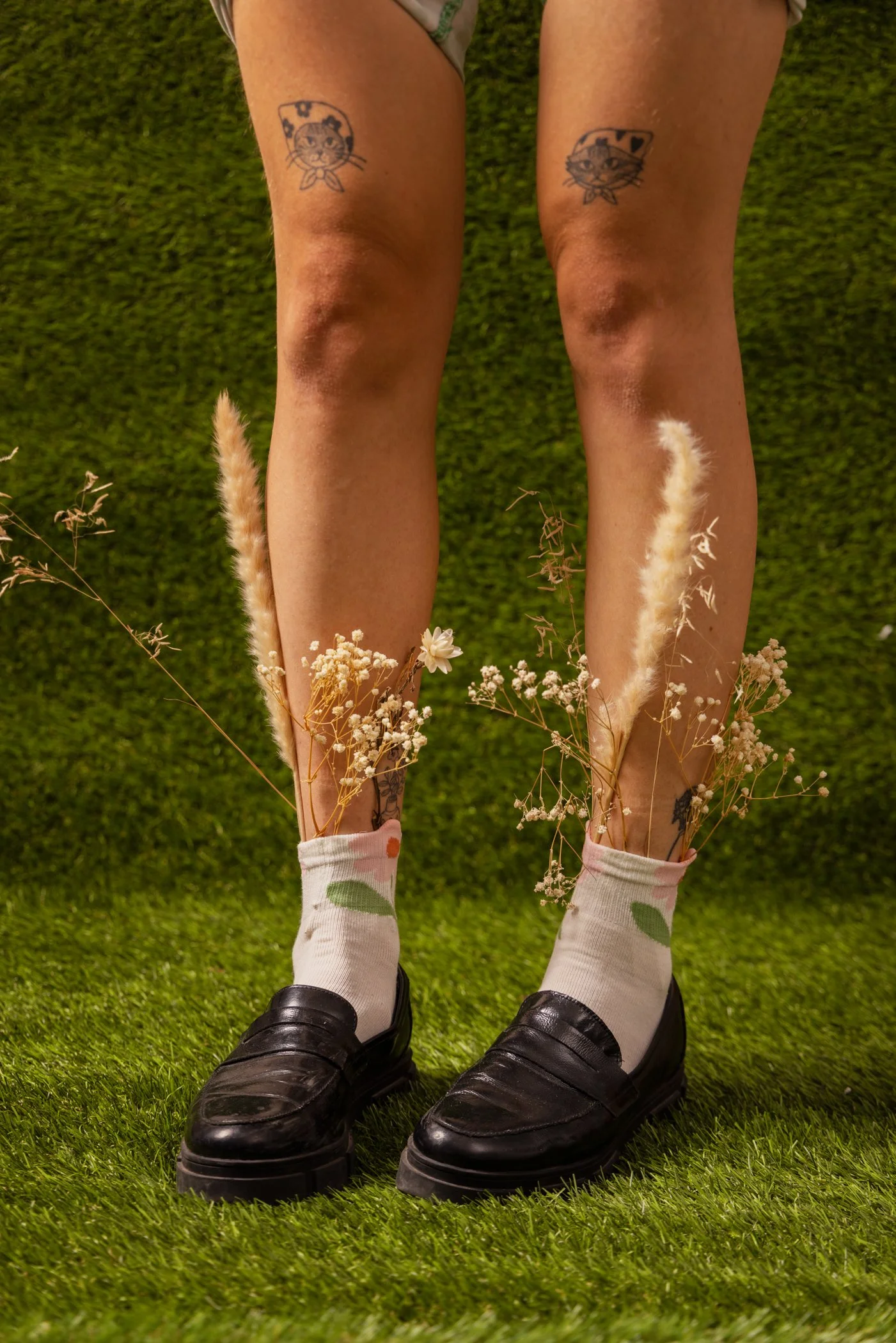 Jambes d'une personne avec des tatouages de chats et des chaussettes à motifs floral, portant des chaussures noires, sur un tapis vert avec des fleurs en premier plan.