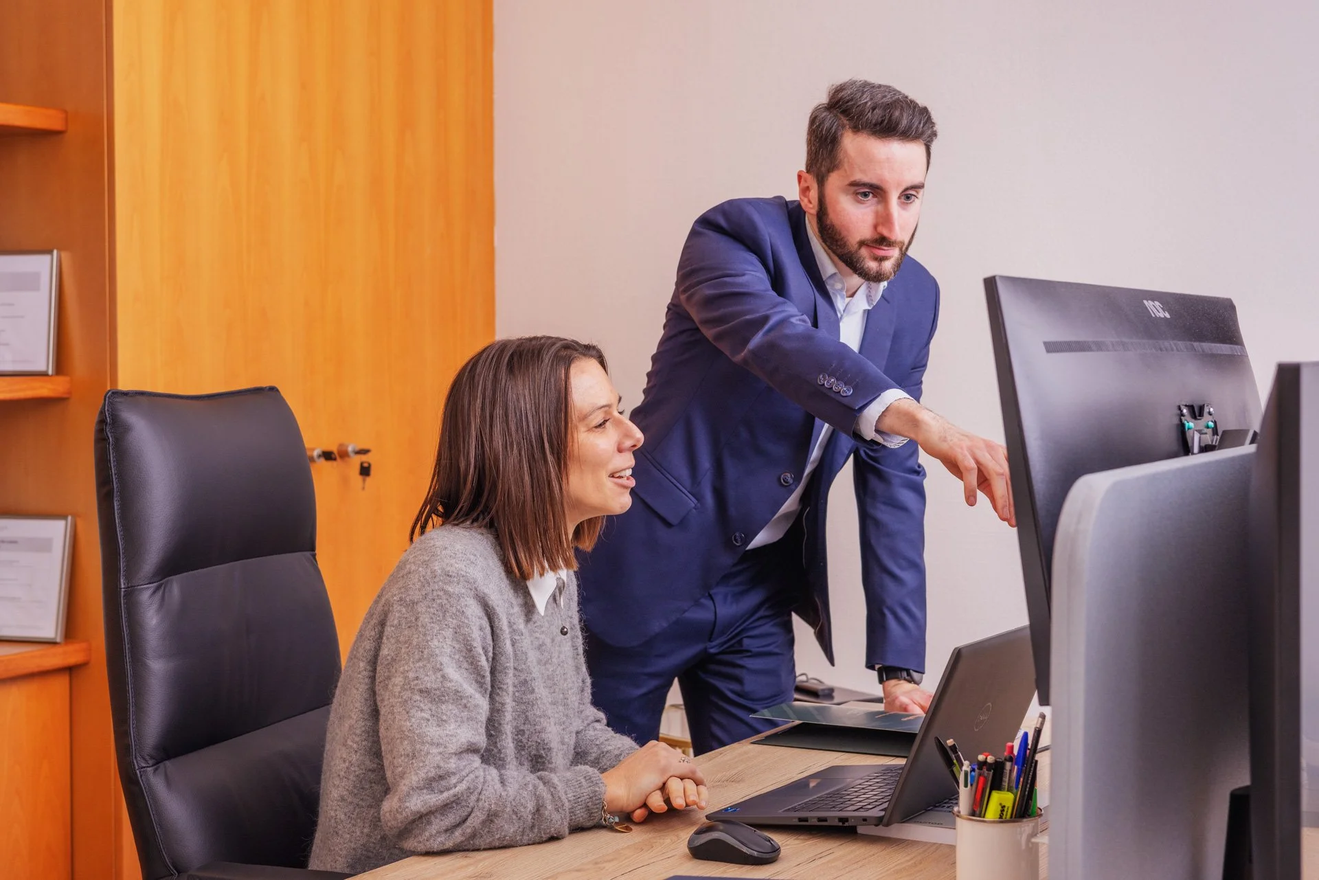 Un homme en costume bleu montre quelque chose à une femme assise à un bureau devant un ordinateur en milieu professionnel.