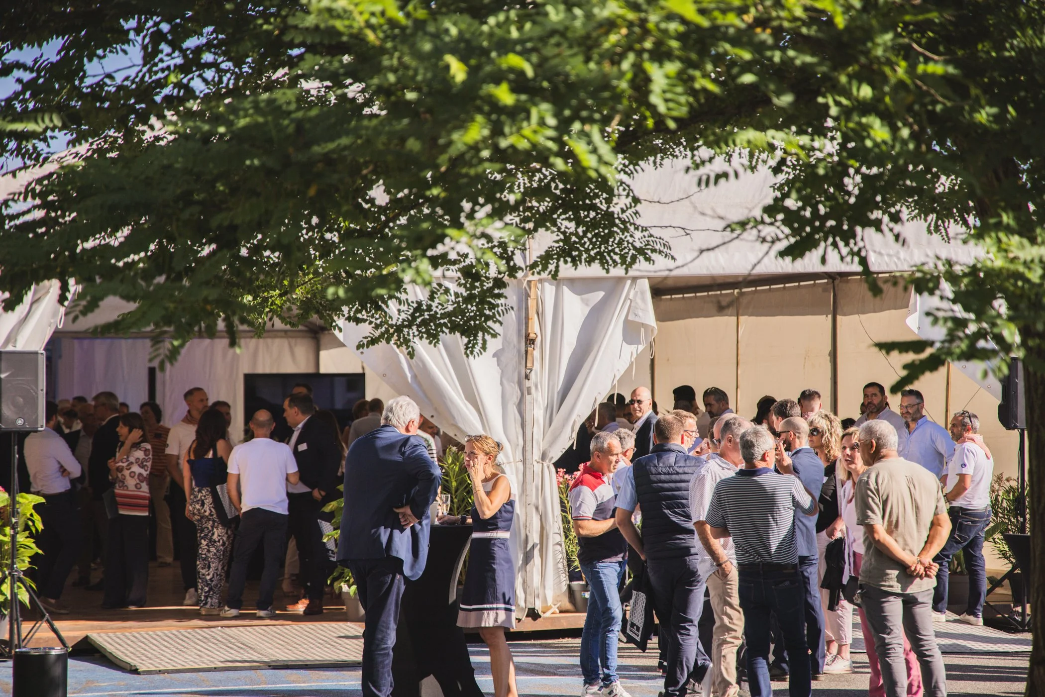 Un grand groupe de personnes assises ou debout sous un chapiteau blanc lors d'un événement en plein air, avec des arbres verts en arrière-plan