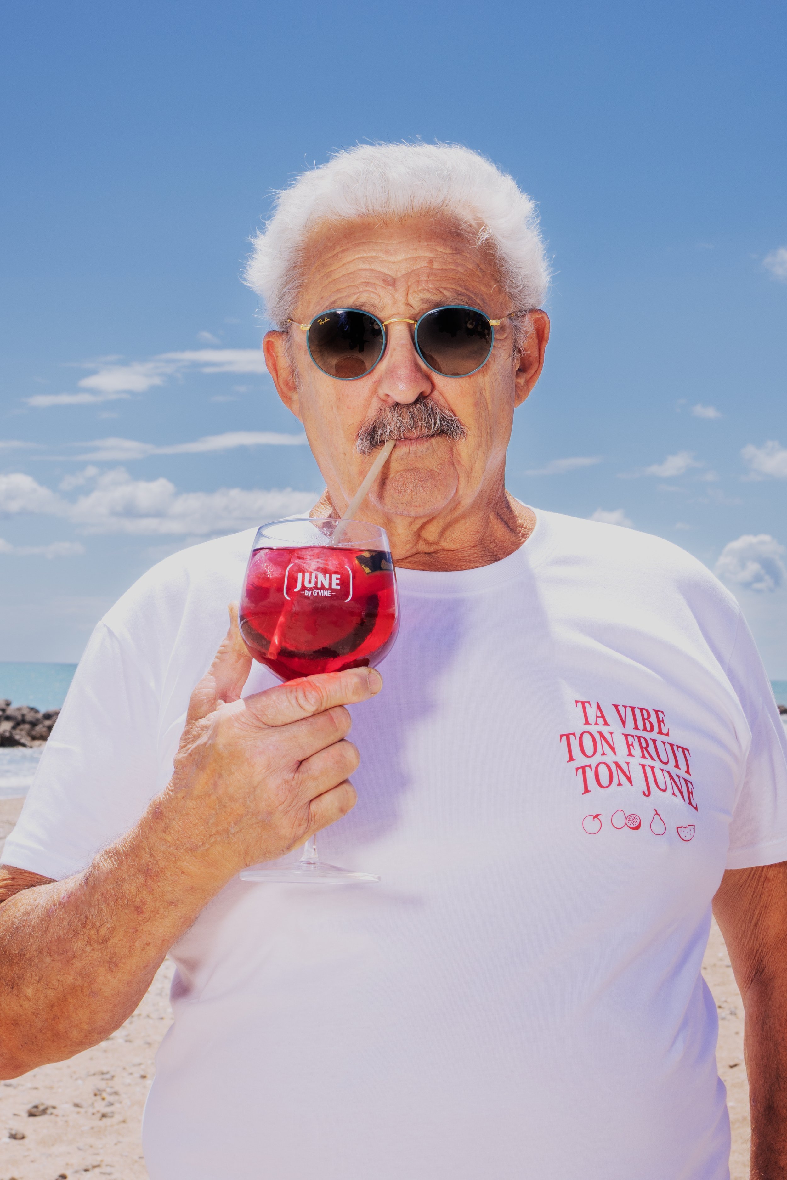 Un homme âgé portant des lunettes de soleil Ray-Ban, une chemise blanche avec un texte rose, tenant un verre de cocktail rouge avec une paille, sur une plage ensoleillée avec un ciel bleu et quelques nuages.