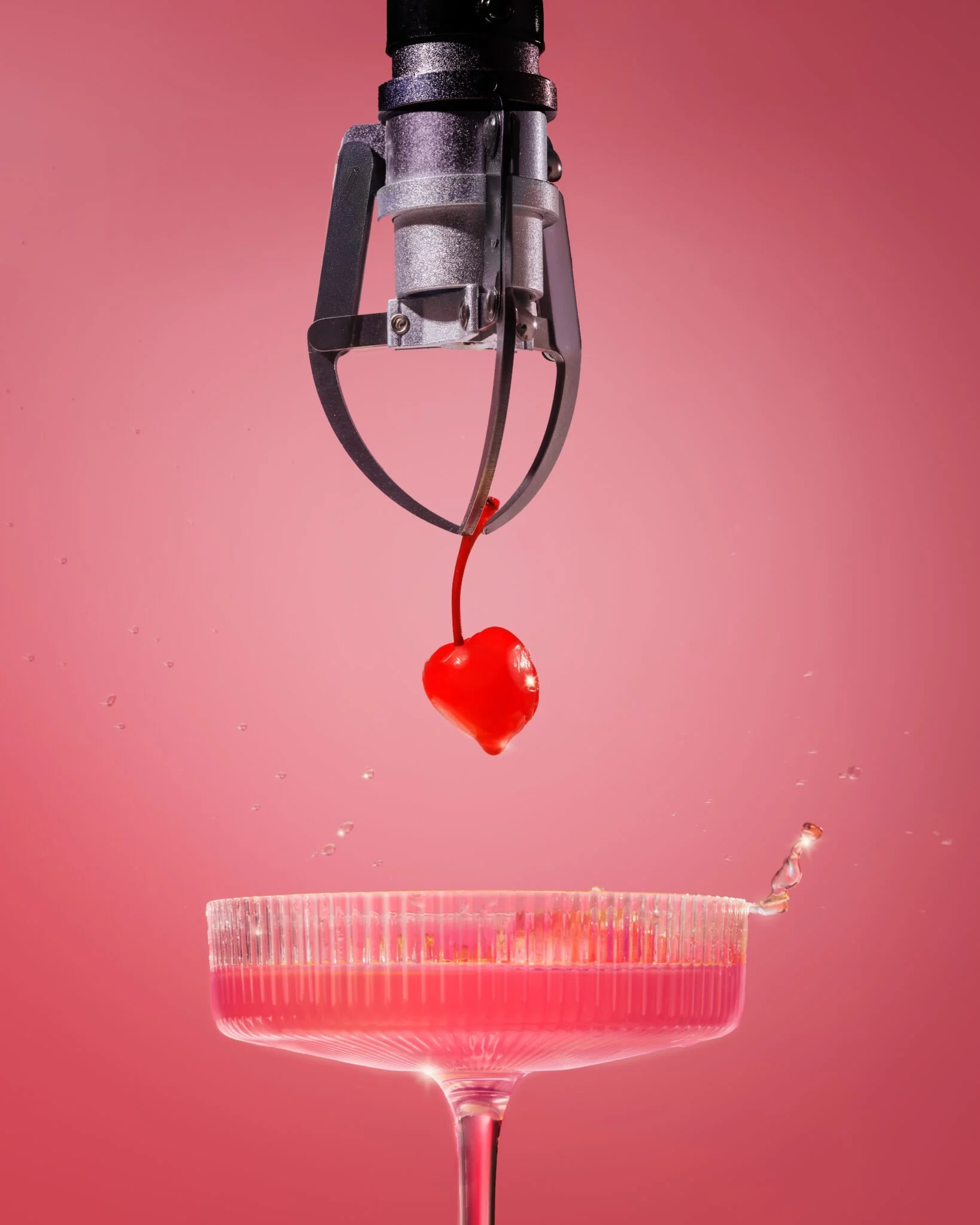 Une cerise rouge tombe dans un verre à cocktail, à l'intérieur du verre se trouve une coupe en papier rose. Un pistolet de tir à ressort est positionné au-dessus de la cerise.