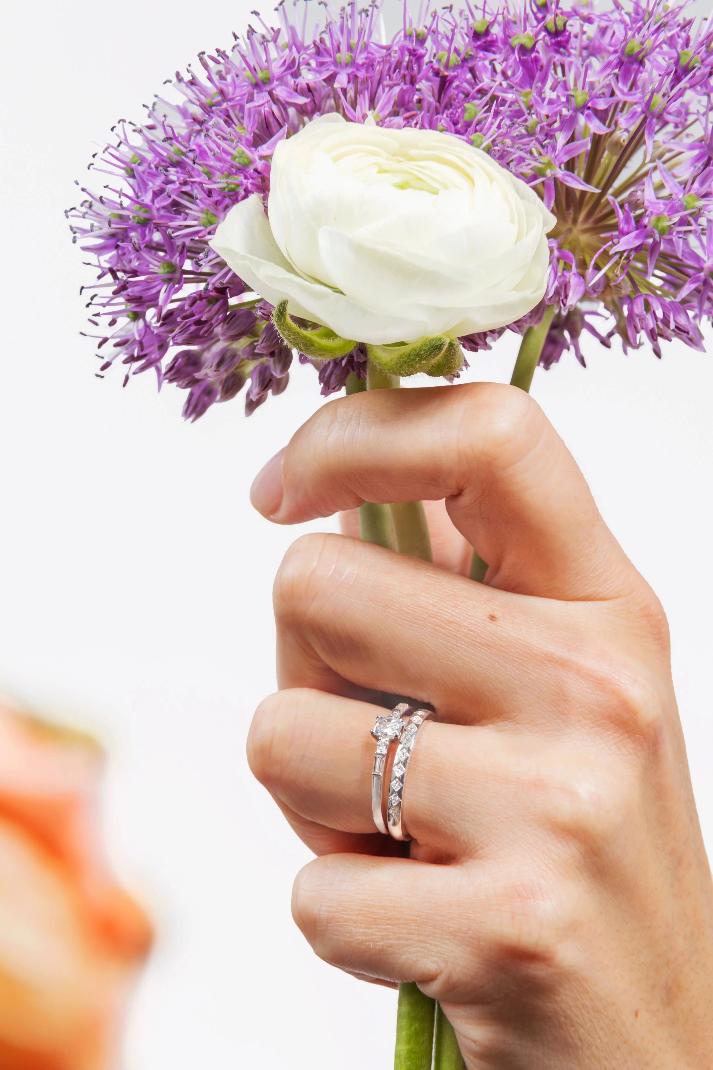 Une main tenant un bouquet de fleurs, comprenant une fleur blanche ressemblant à une rose et une grande fleur violette, avec des bagues en argent à l'annulaire.