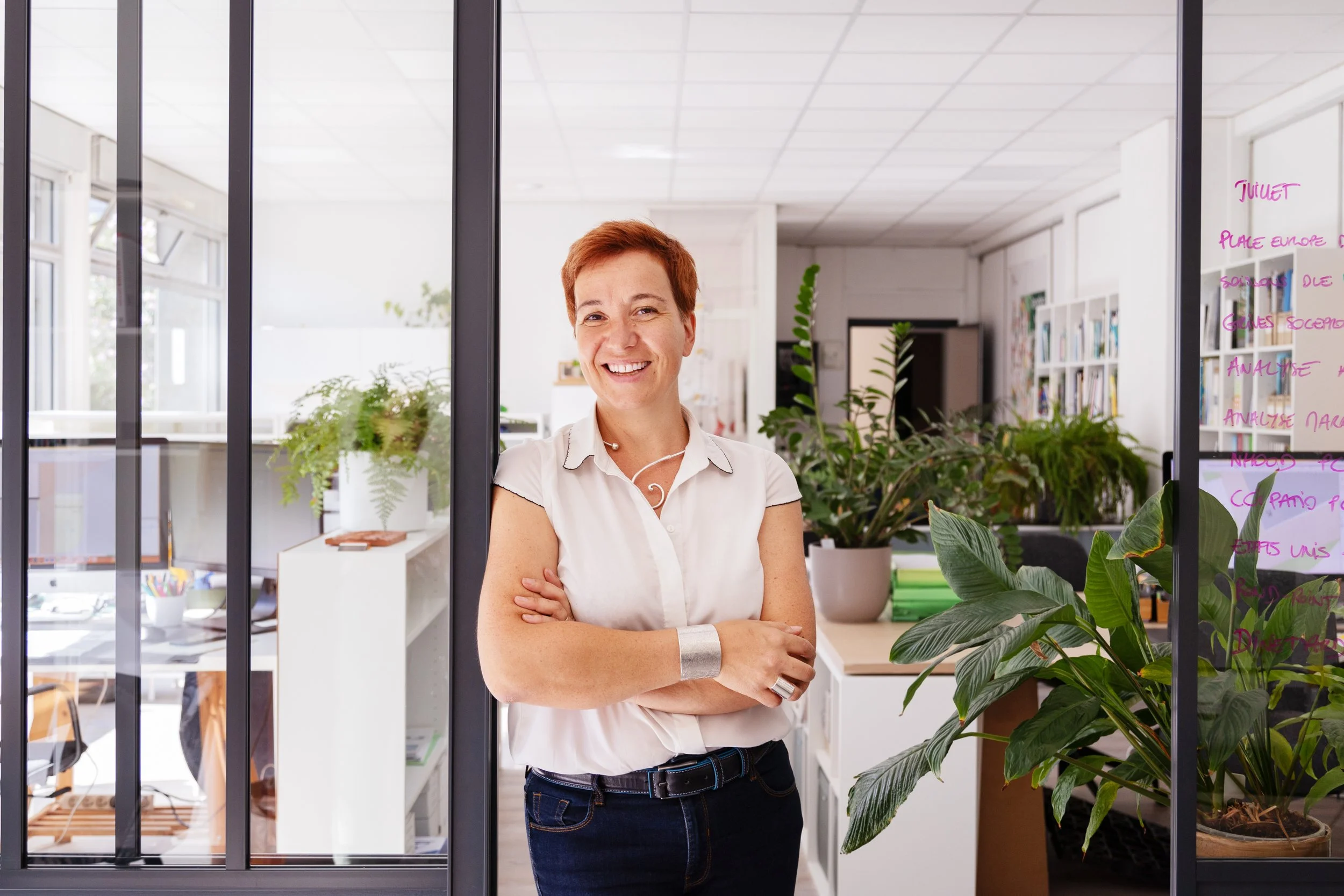 Femme souriante avec vêtements décontractés, debout dans un espace de bureau moderne, derrière une grande fenêtre en verre, entourée de plantes vertes et de livres, avec écriture en rose sur la vitre.