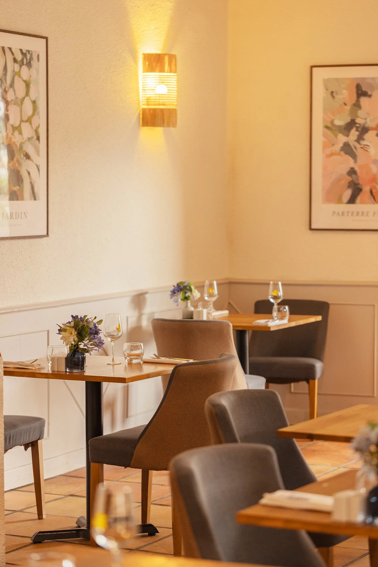 Salle à manger d'un restaurant avec des tables en bois, des chaises en tissu gris, des fleurs violettes sur les tables et des tableaux abstraits au mur, éclairée par une applique chaleureuse.