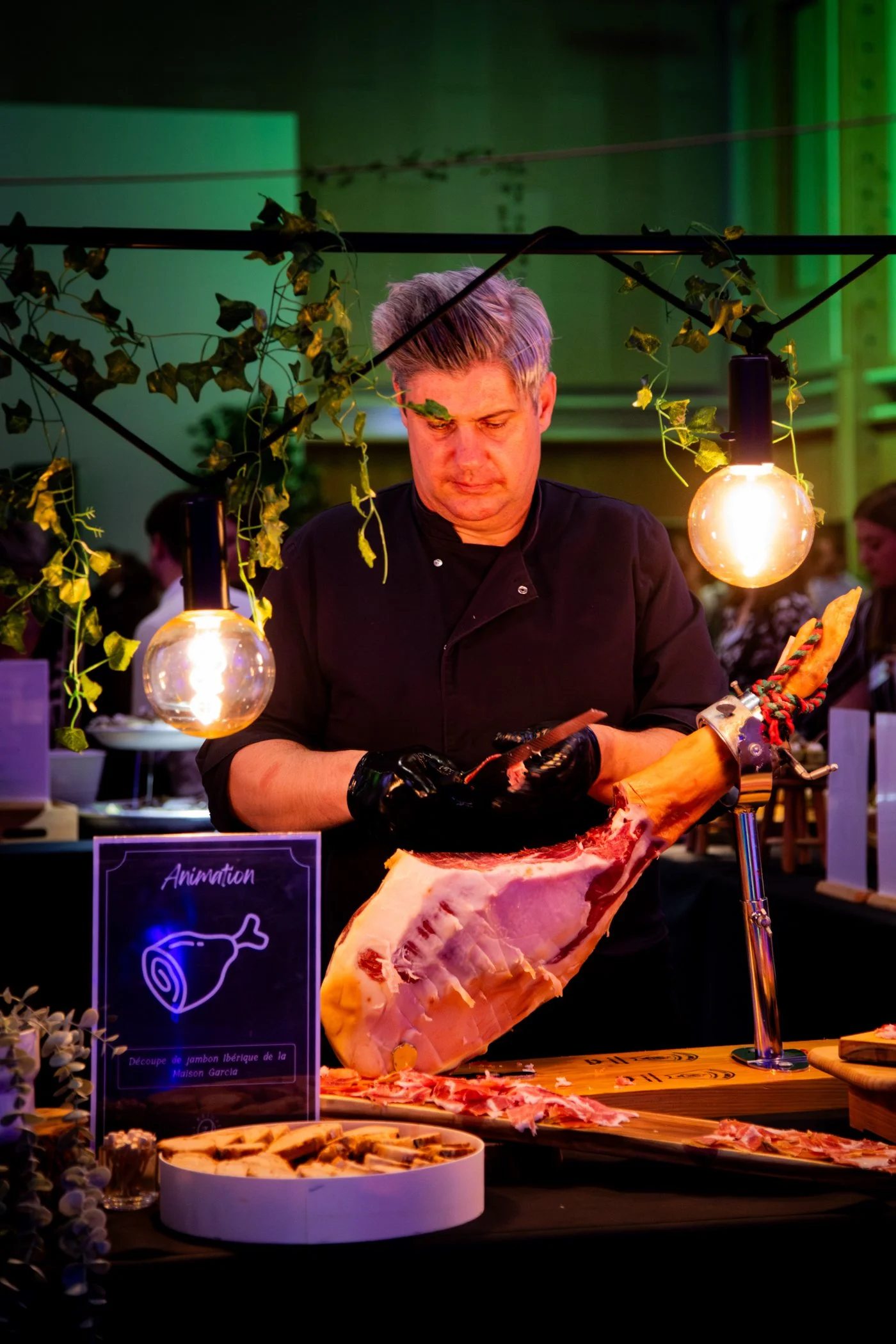 Un chef préparant un morceau de jambon ibérique lors d'un événement, avec une présentation artistique de la viande et des décorations de lumières et de plantes.