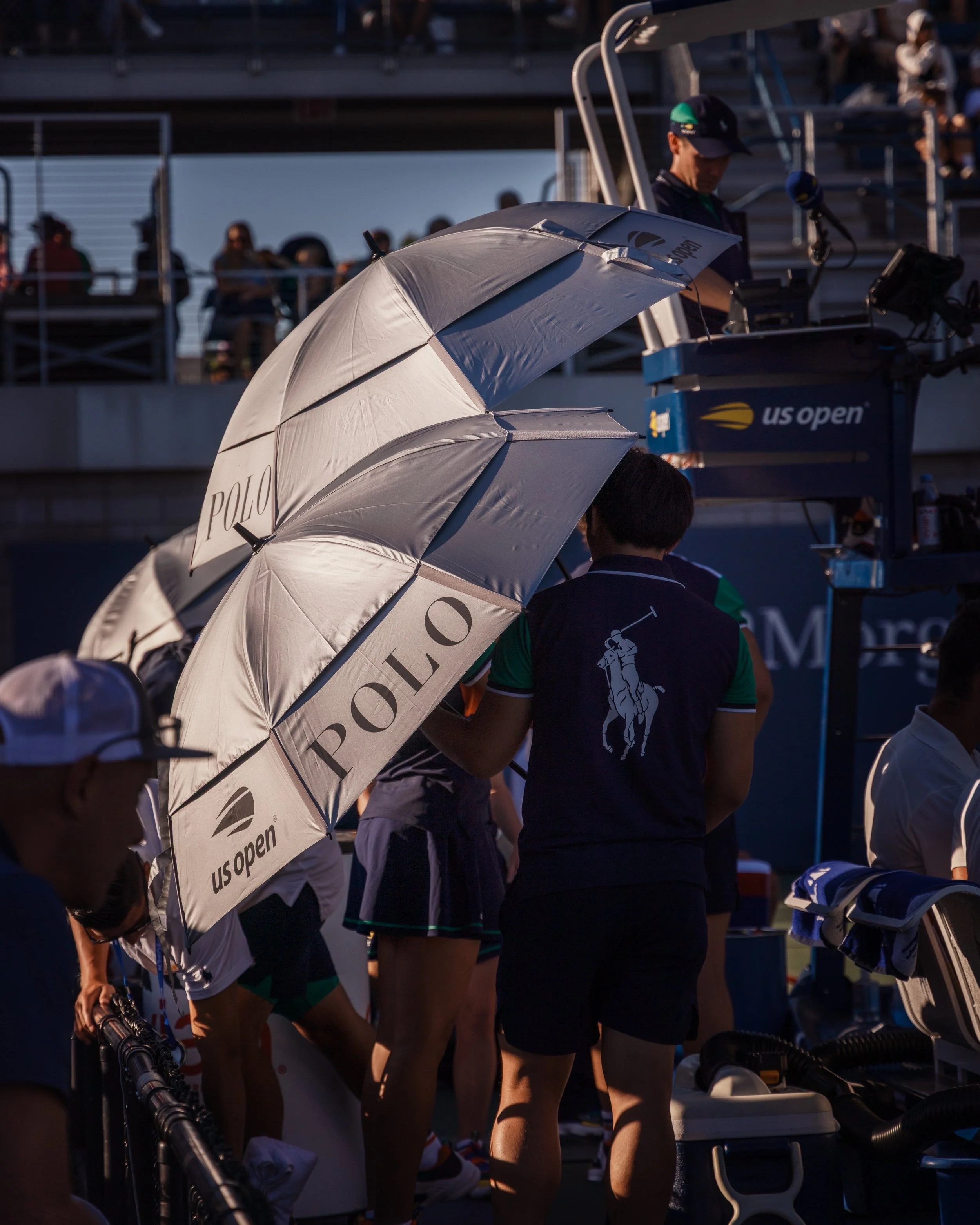 Une scène de l'US Open de tennis, avec plusieurs joueuses debout, en uniforme sportif, sous un parasol blanc portant le logo 'Polo'. Des spectateurs sont visibles en arrière-plan, assis dans les gradins et debout, certains avec des parasols ou des ca