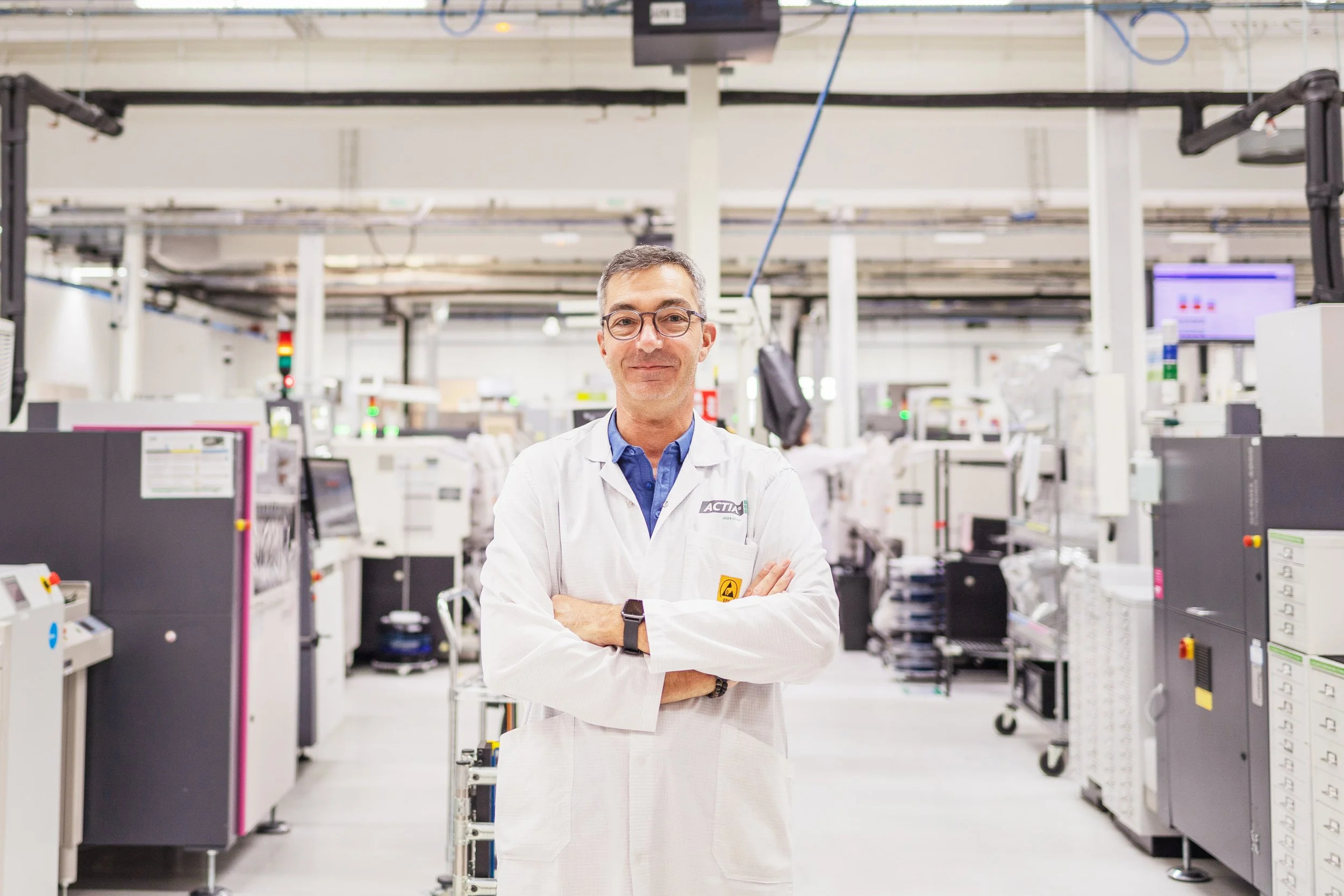 Un homme souriant, portant des lunettes, un manteau blanc et une montre, se tient dans une usine ou un centre de traitement industriel avec des équipements et des machines électroniques en arrière-plan.