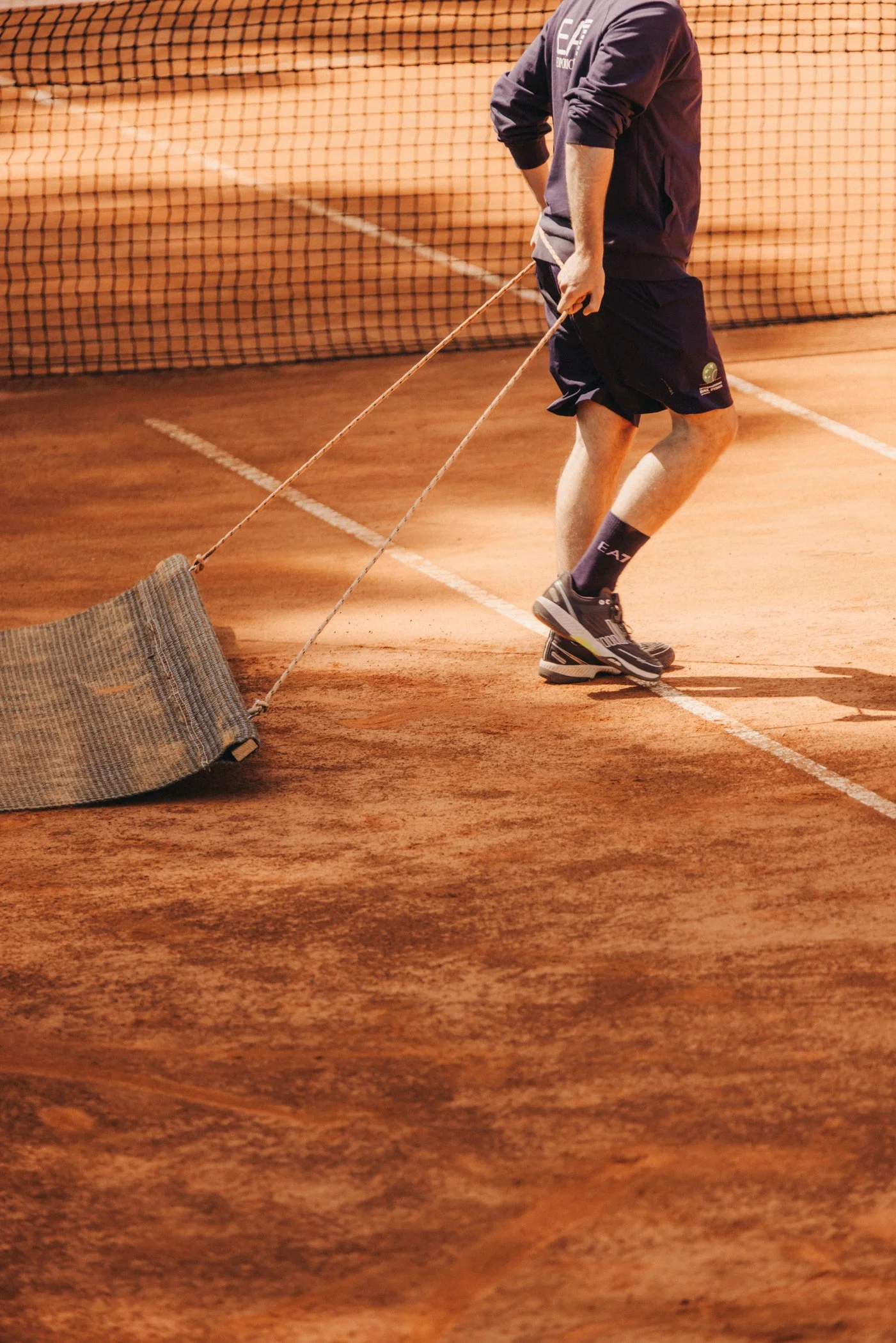 Une personne joue au tennis sur un court en terre battue, portant une veste mauve, un short noir et des chaussures de sport, avec une haie ou des grilles de clôture en arrière-plan.