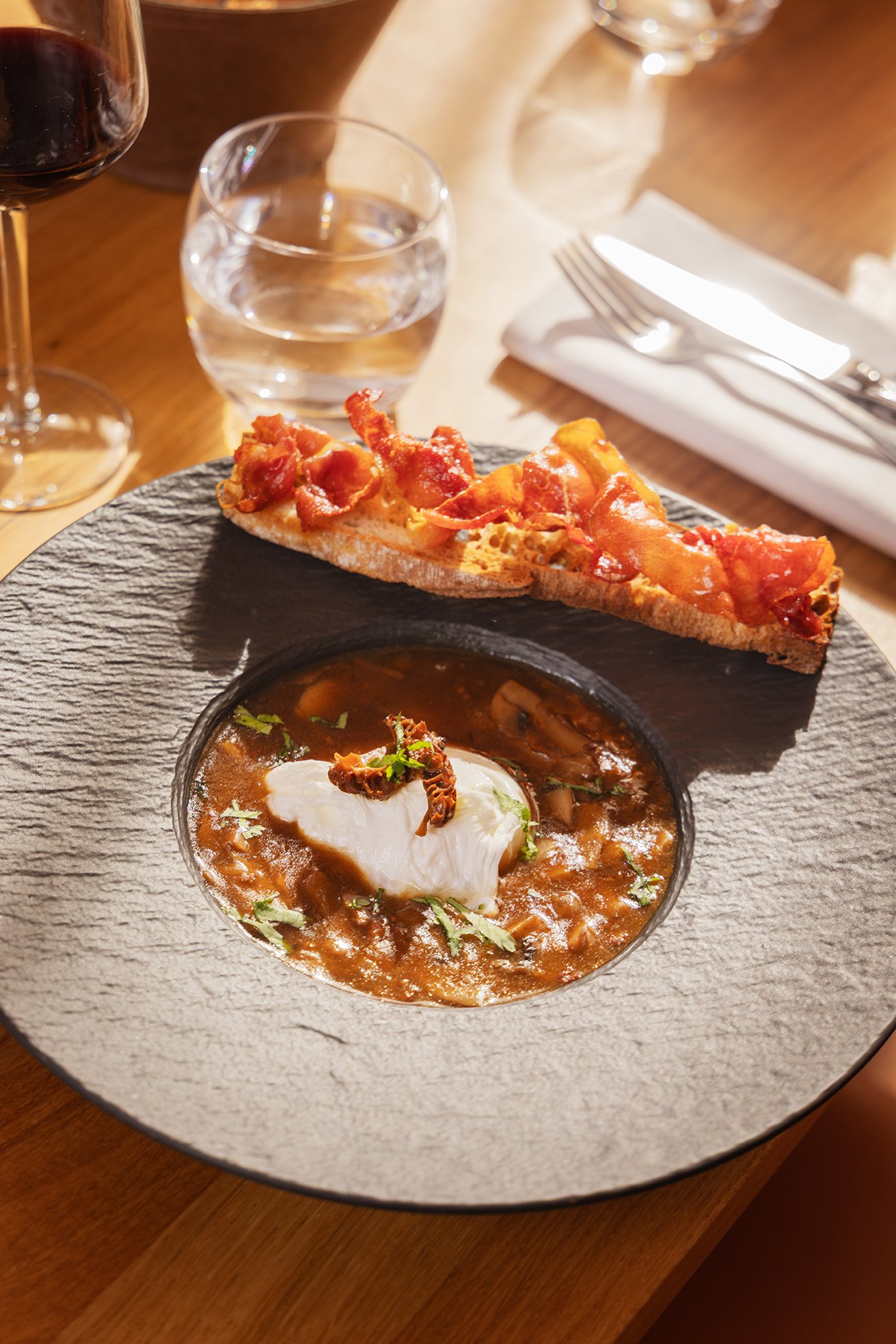 Soupe à la viande, œuf poché et garniture dans un bol en céramique, accompagné d'une tranche de pizza bacon, verre d'eau, verre de vin rouge, fourchette, couteau et serviette en papier sur une table en bois.