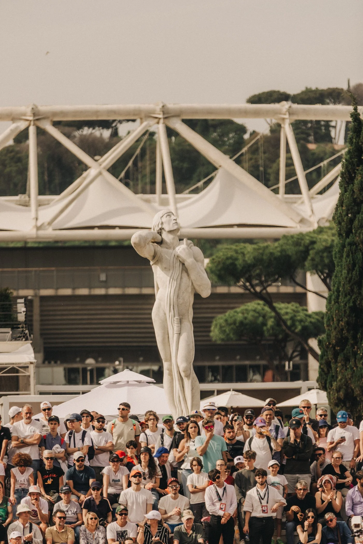 Une statue représentant une femme de style Art déco tenant une colonne, située dans une foule de personnes assises ou debout lors d'un rassemblement ou d'un événement en plein air.