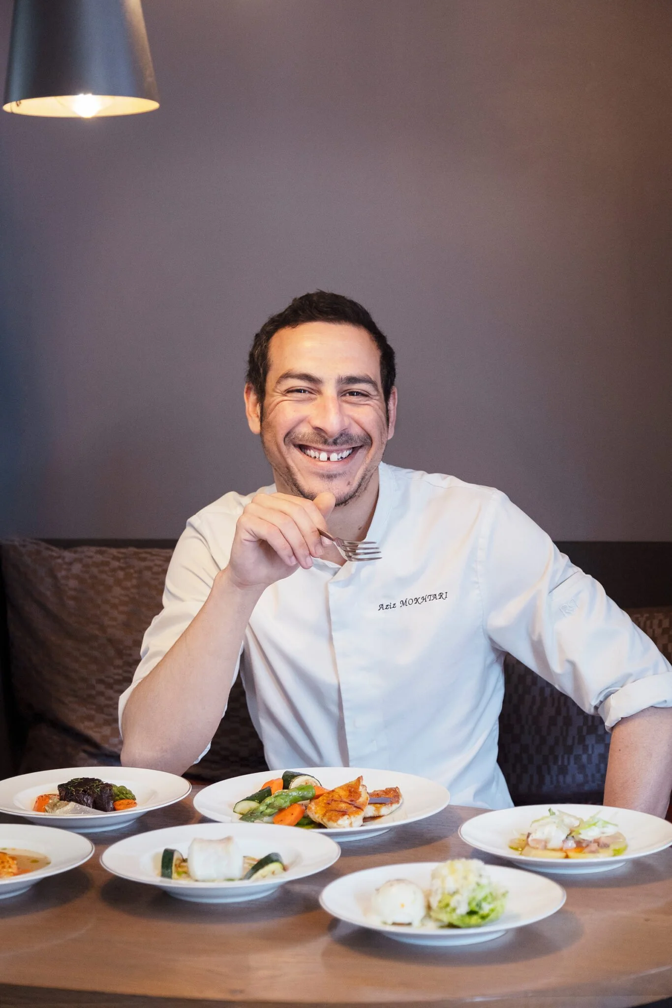 Un homme souriant, portant une veste blanche avec le nom 'Aziz Mokhtari' brodé, est assis à une table de restaurant avec plusieurs plats de nourriture en face. Il tient une fourchette dans sa main droite.