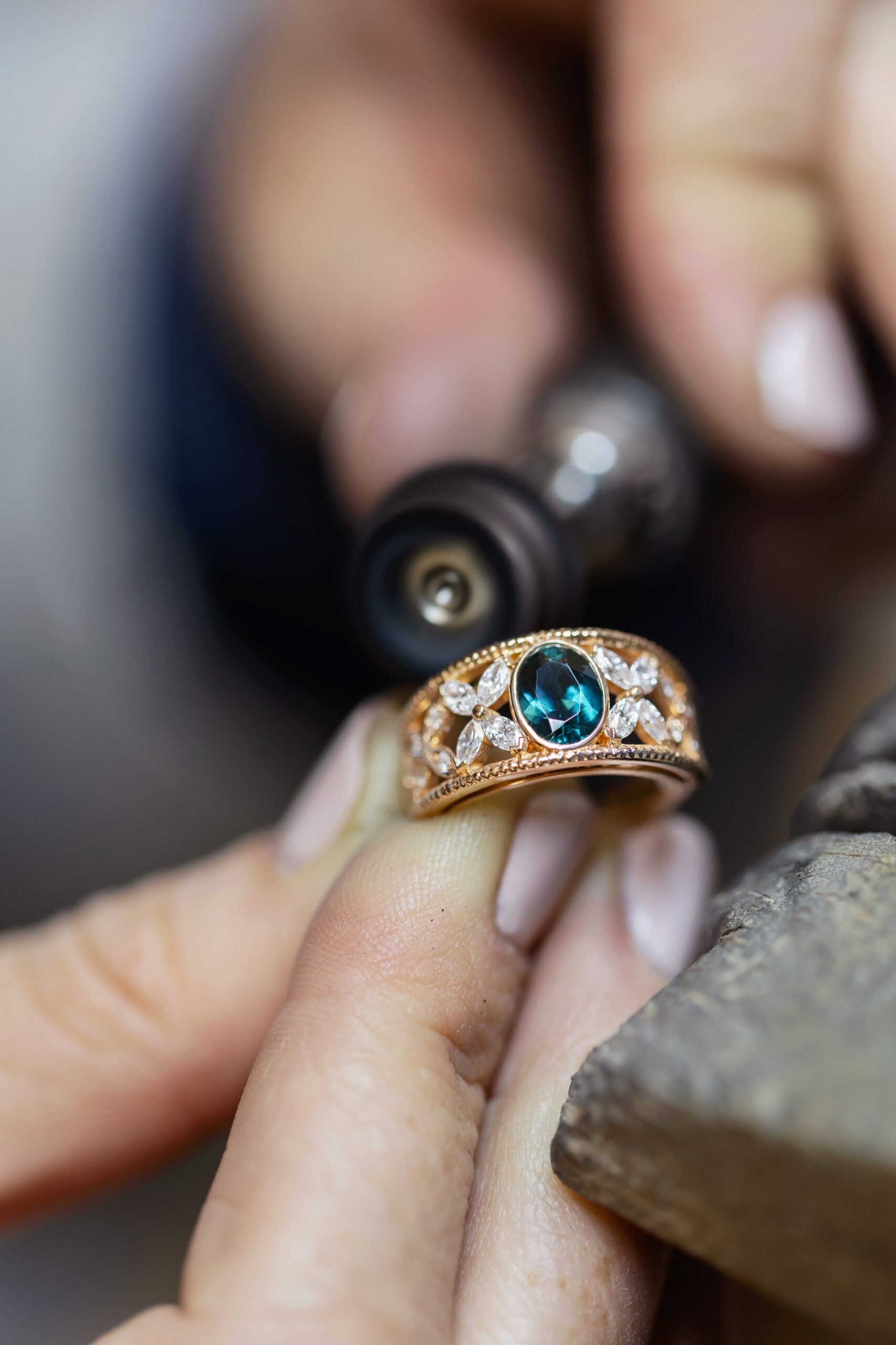 Une main portant une bague en or avec un grand saphir bleu entouré de diamants, tenue contre une surface rocheuse.
