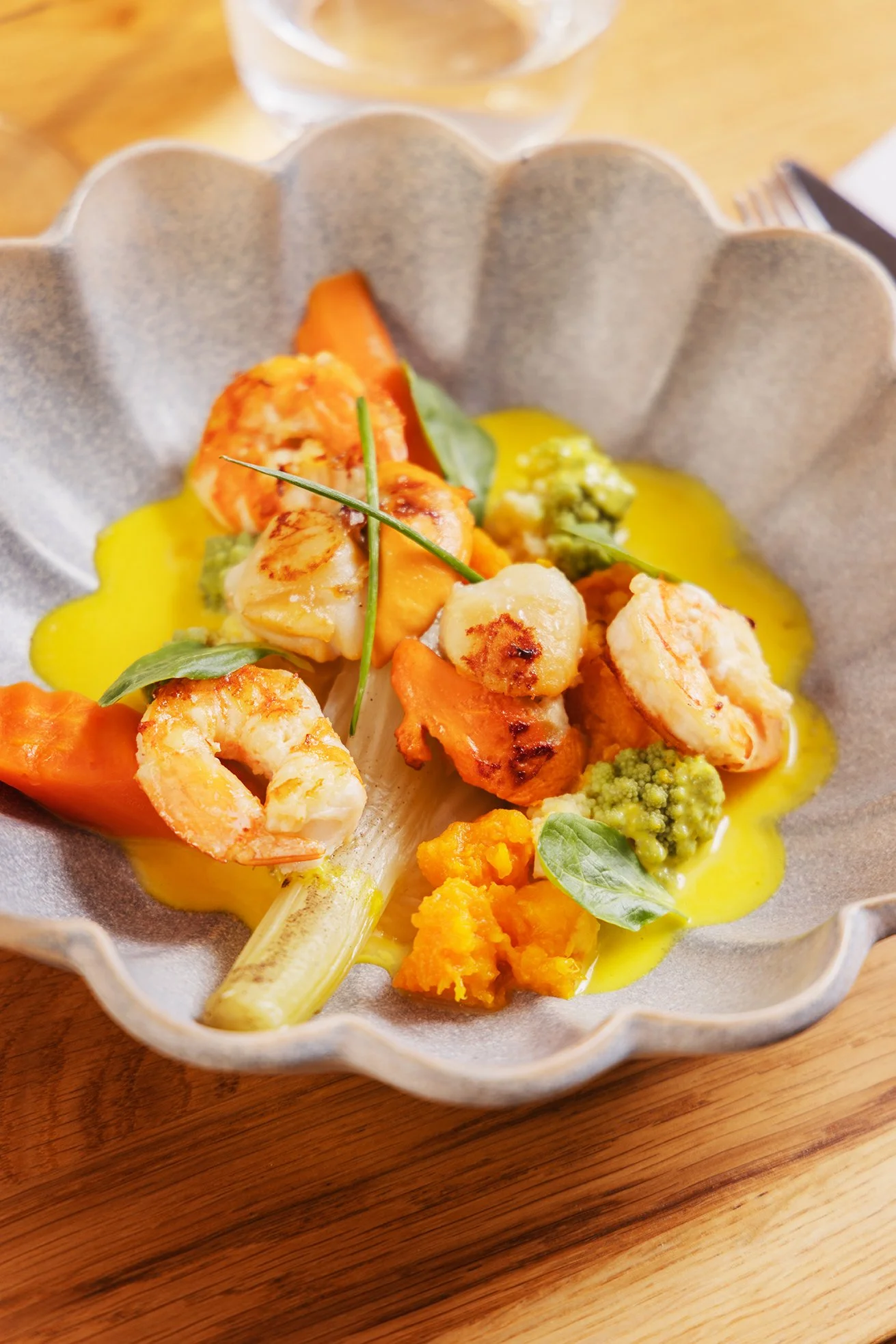 Assiette creuse avec crevettes, légumes colorés, sauce jaune, et herbes, posée sur une table en bois.