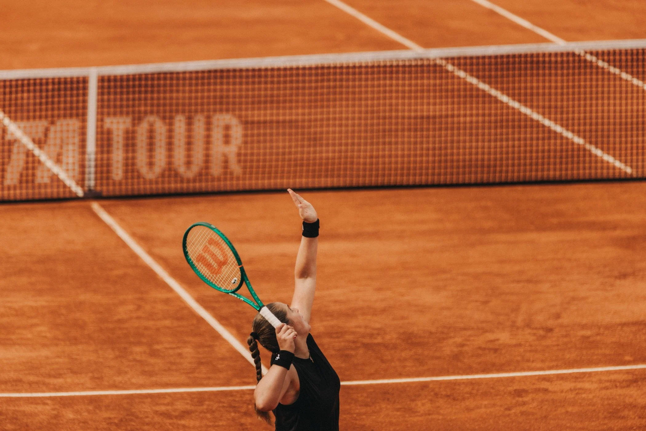 Une joueuse de tennis en noir, vue de dos, s'apprête à servir lors d'un match sur un court en terre battue.