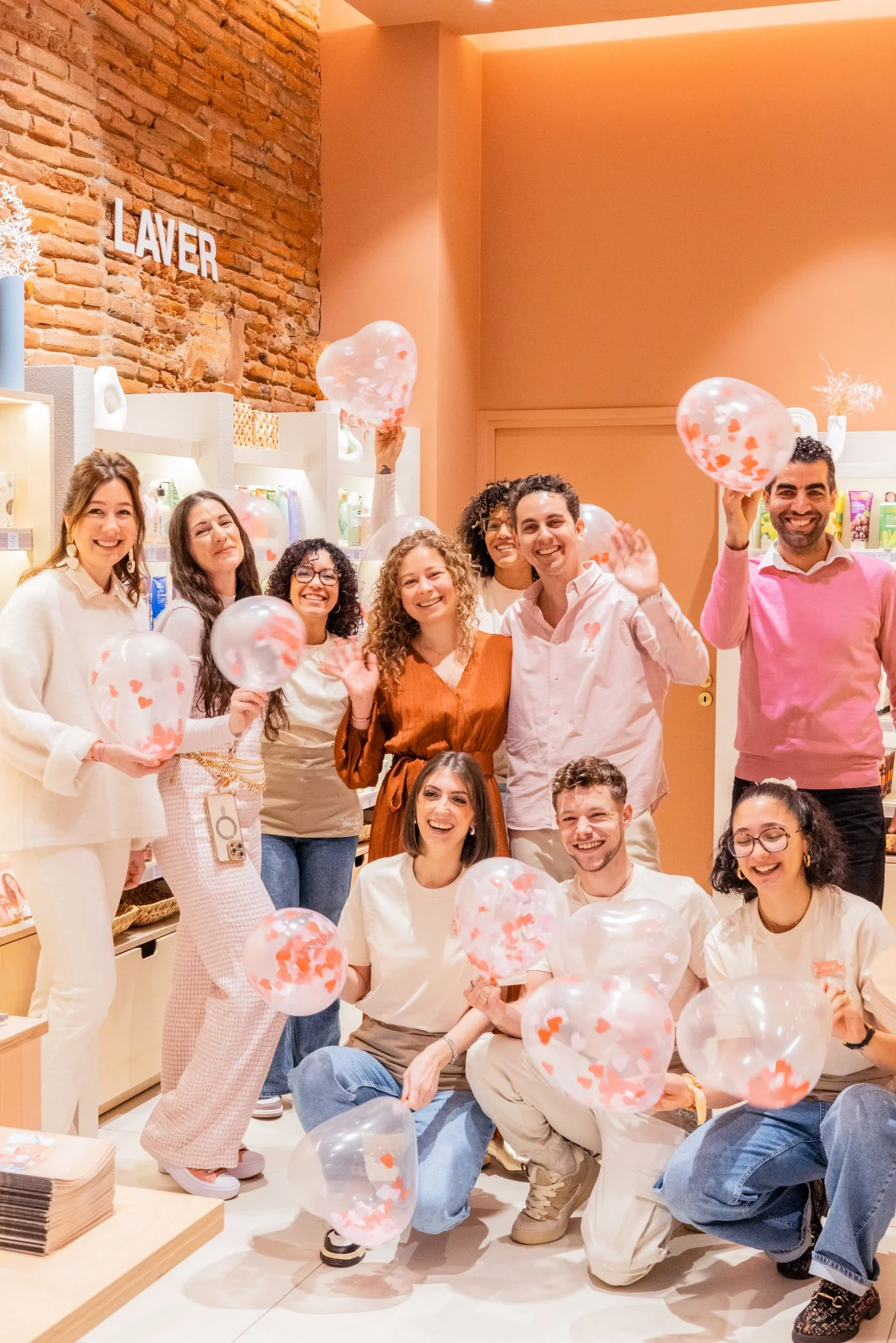 Groupe de personnes souriantes tenant des ballons en plastique avec des cœurs orange et blancs, dans une boutique ou un espace intérieur. Mur en briques et décor lumineux en arrière-plan.