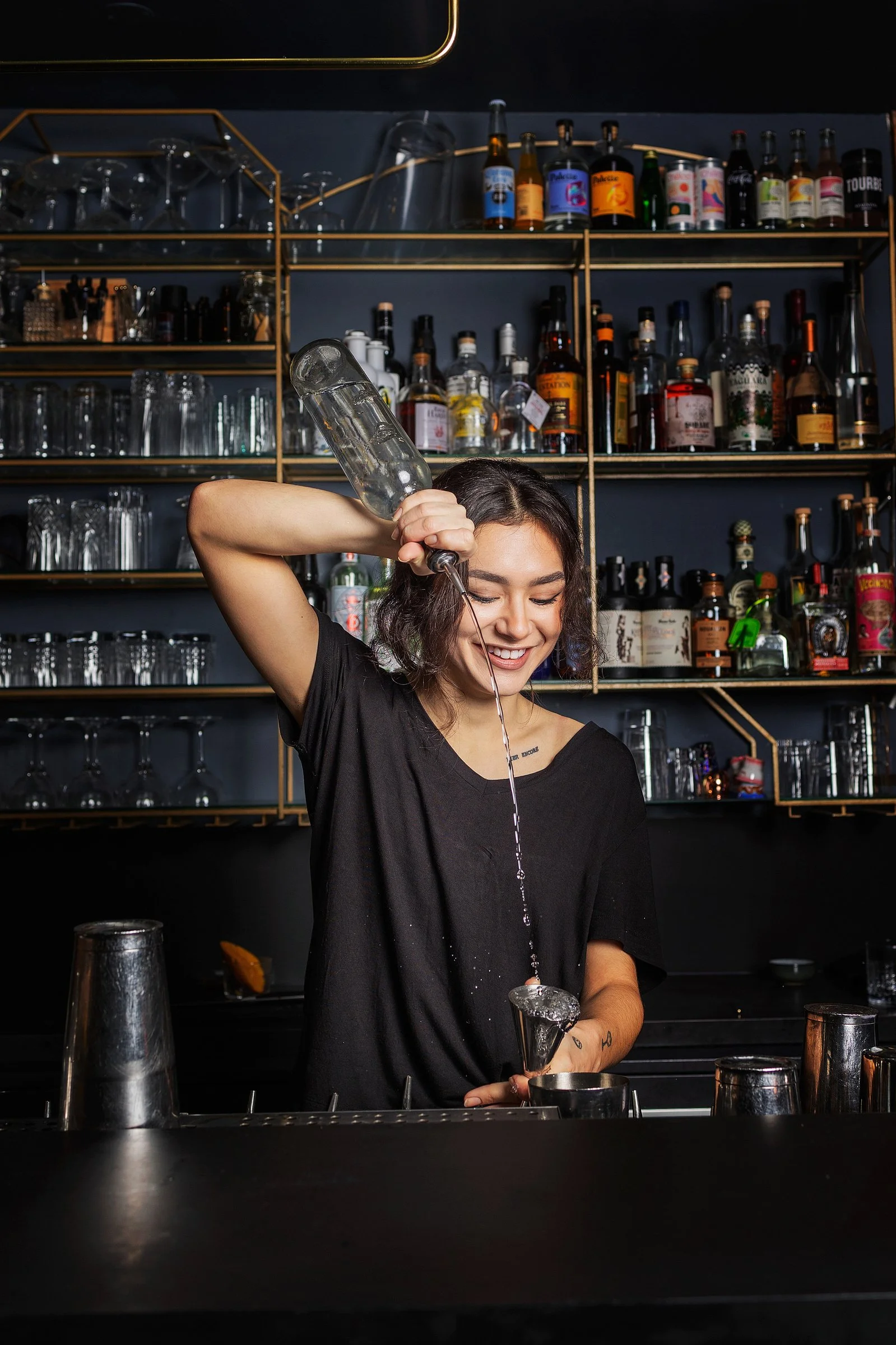 Une jeune femme souriante en noir, versant de l'alcool dans un verre à cocktail derrière le bar, avec des étagères remplies de bouteilles d'alcool en arrière-plan.