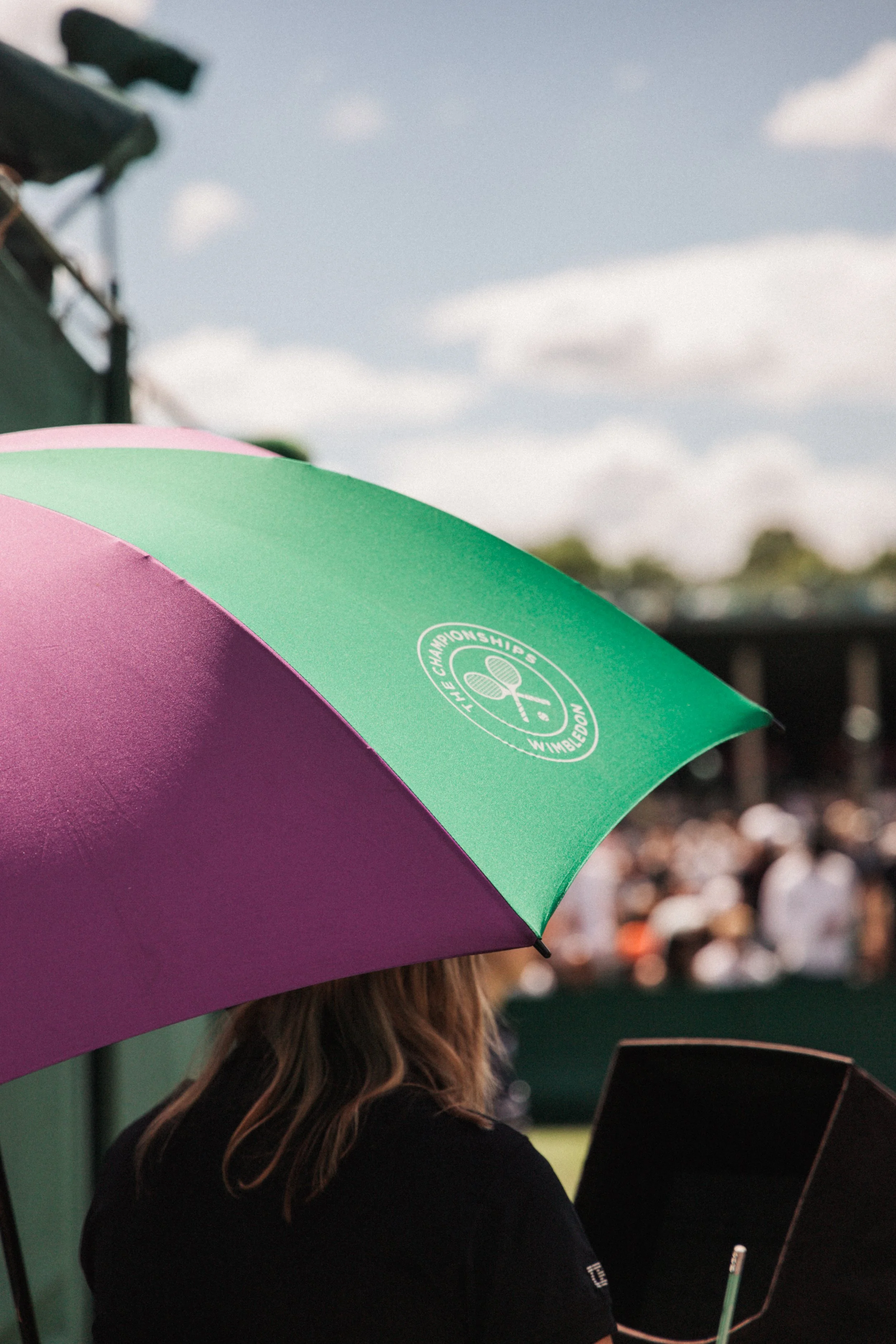 Une femme avec des cheveux bruns portant un t-shirt noir et tenant un parapluie rose et vert lors d'un événement en plein air à Wimbledon.