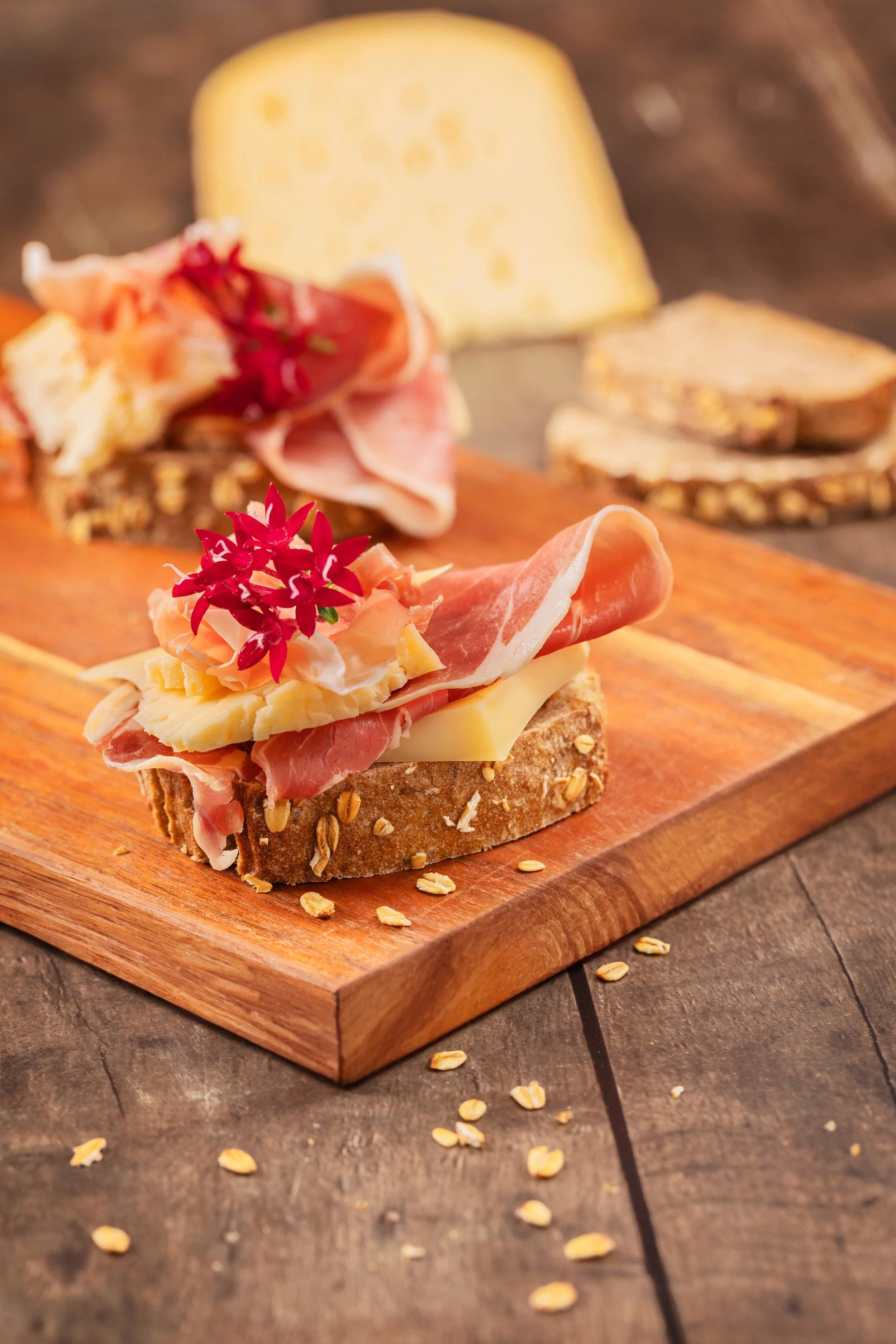 Tartine de pain avec fromage, jambon et fleurs roses, sur une planche en bois, avec des graines d'avoine.