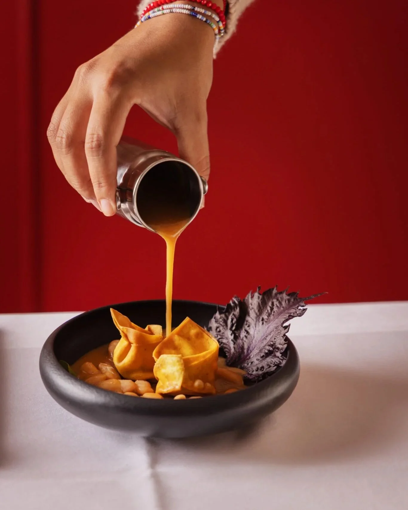 Une main verse une sauce brune dans un bol contenant des chips et une salade, avec une feuille de salade violette sur le côté, fond rouge.