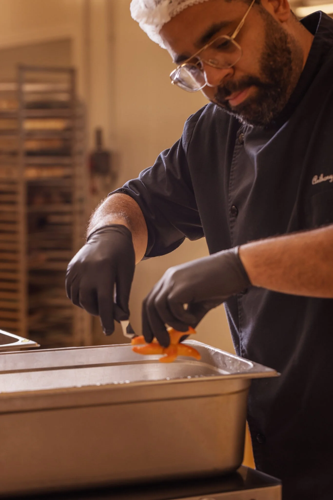 Un homme portant un chapeau de douche, des lunettes, un tablier noir et des gants noirs prépare quelque chose dans une cuisine, probablement en train de cuisiner.