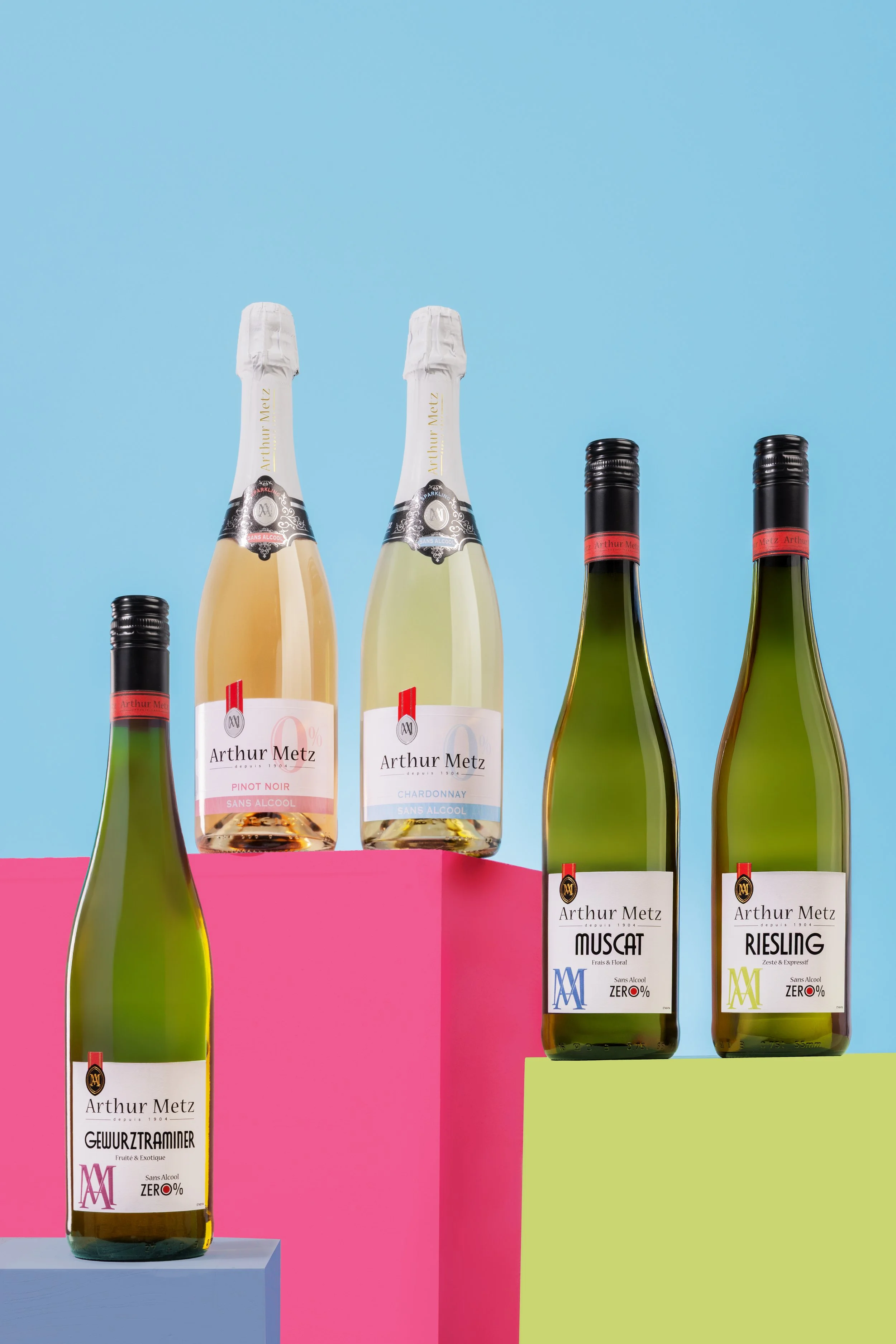 Gamme de bouteilles de vin sans alcool Arthur Metz sur des piédestaux colorés, fond bleu avec accents rose et vert.