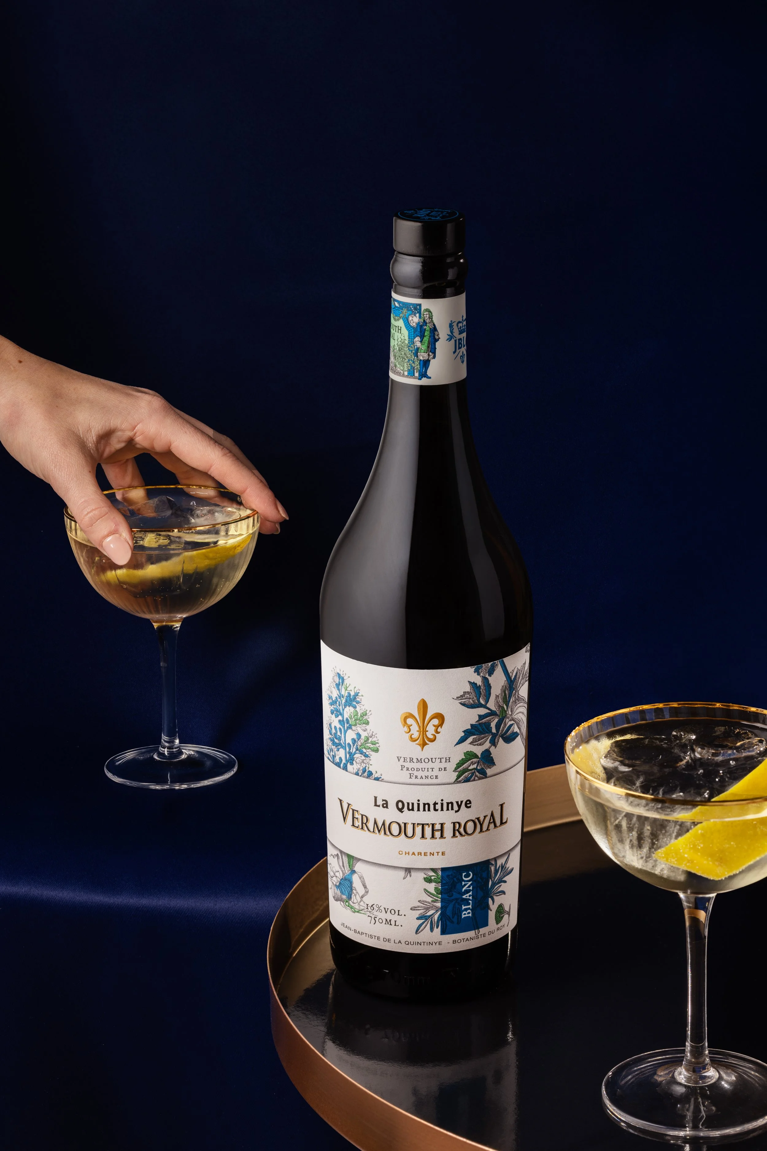 Une bouteille de vermouth La Quintinye Vermouth Royal posée sur une surface ronde en cuivre, entourée de deux verres à cocktail contenant du vermouth avec des tranches de citron, le tout avec un fond sombre.